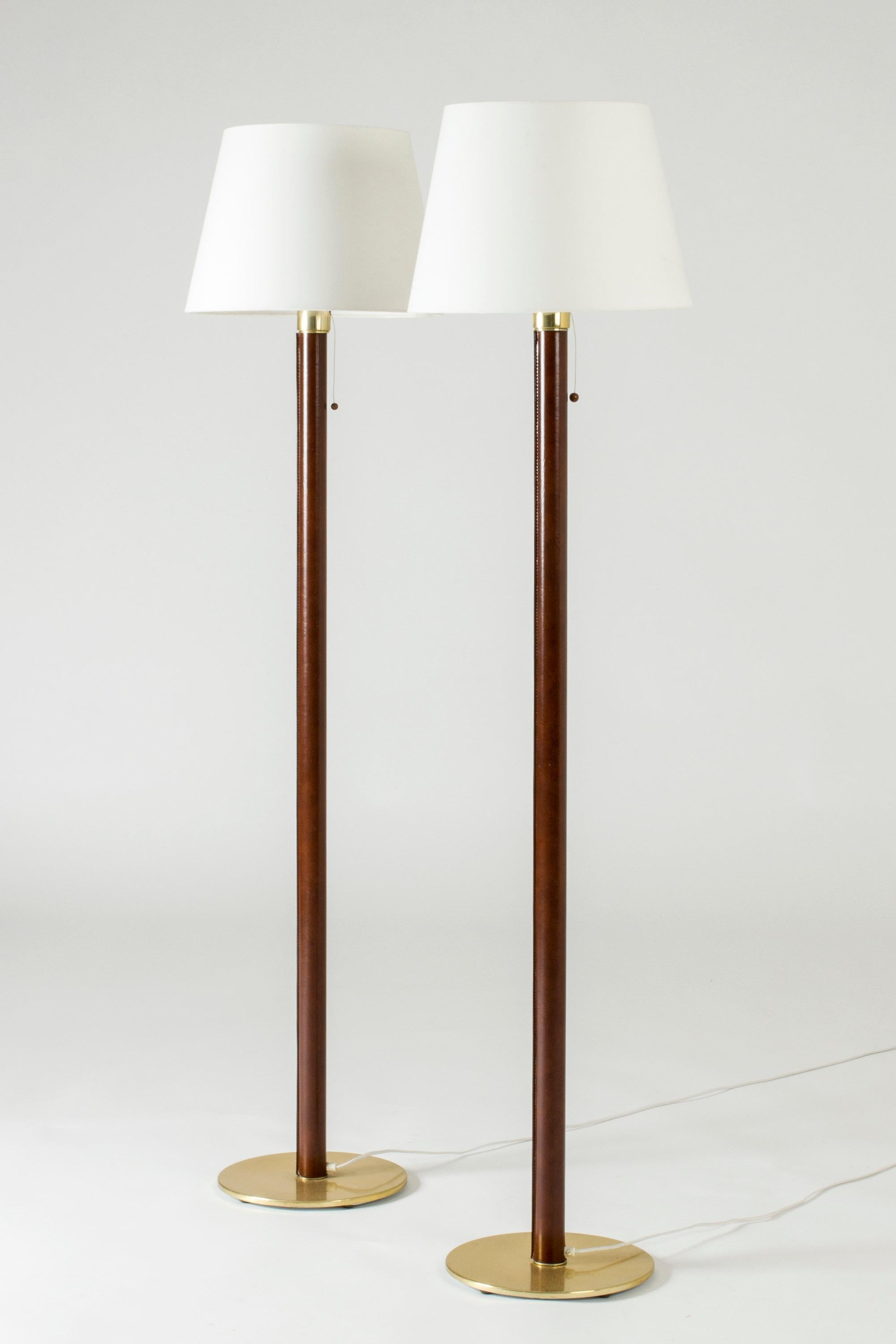 Pair of elegant floor lamps from Falkenbergs Belysning, made from brass. Stems dressed with brown faux leather, visible decorative white seams. Drawstring light switches with balls at the ends.