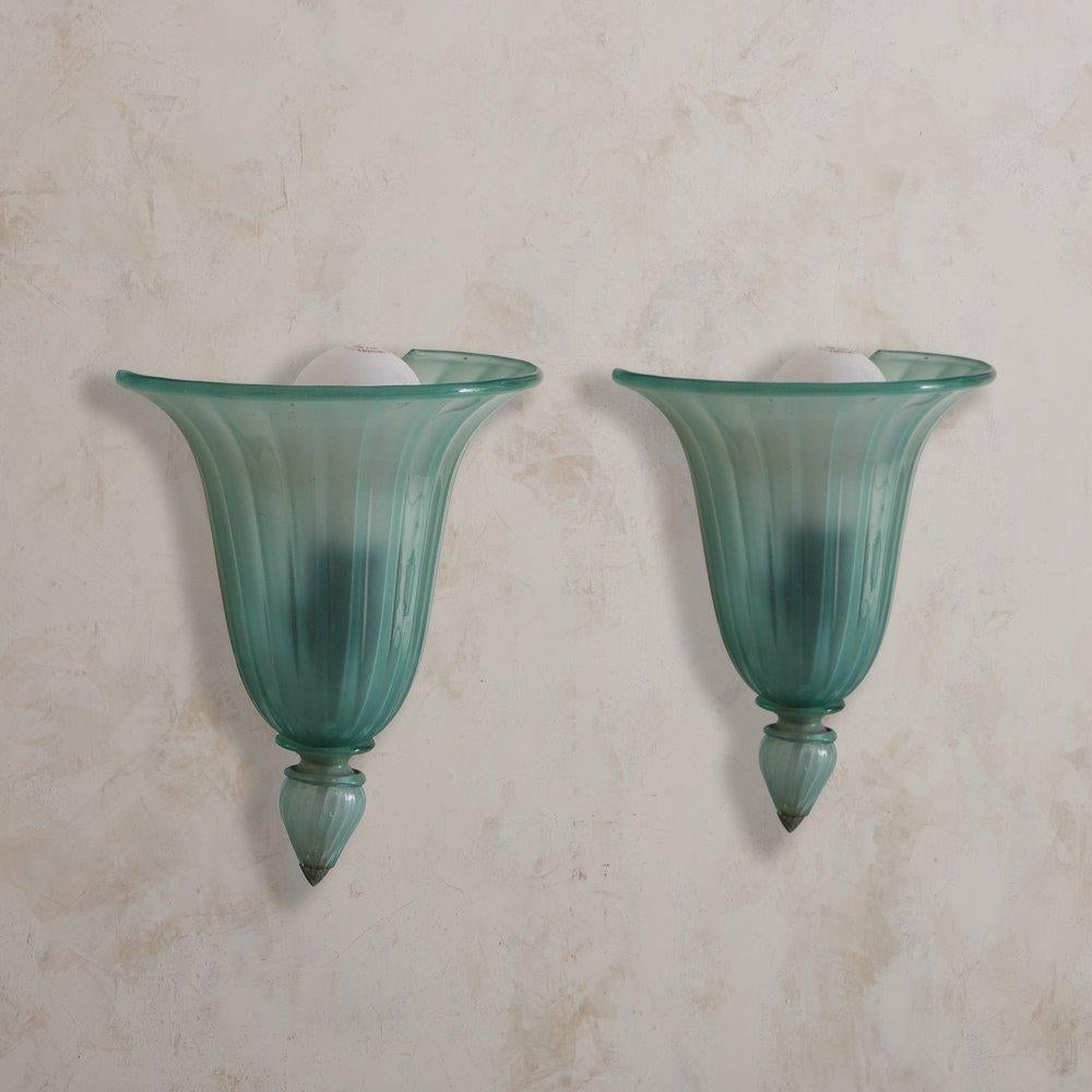 A pair of 1920s hand blown Murano glass wall sconces featuring shell-shaped tapered glass shades in a gorgeous blue hue. Each shade has fluted detailing, a flared demilune top and a bottom finial with a silver metal tip. Unmarked. Sourced in Italy,