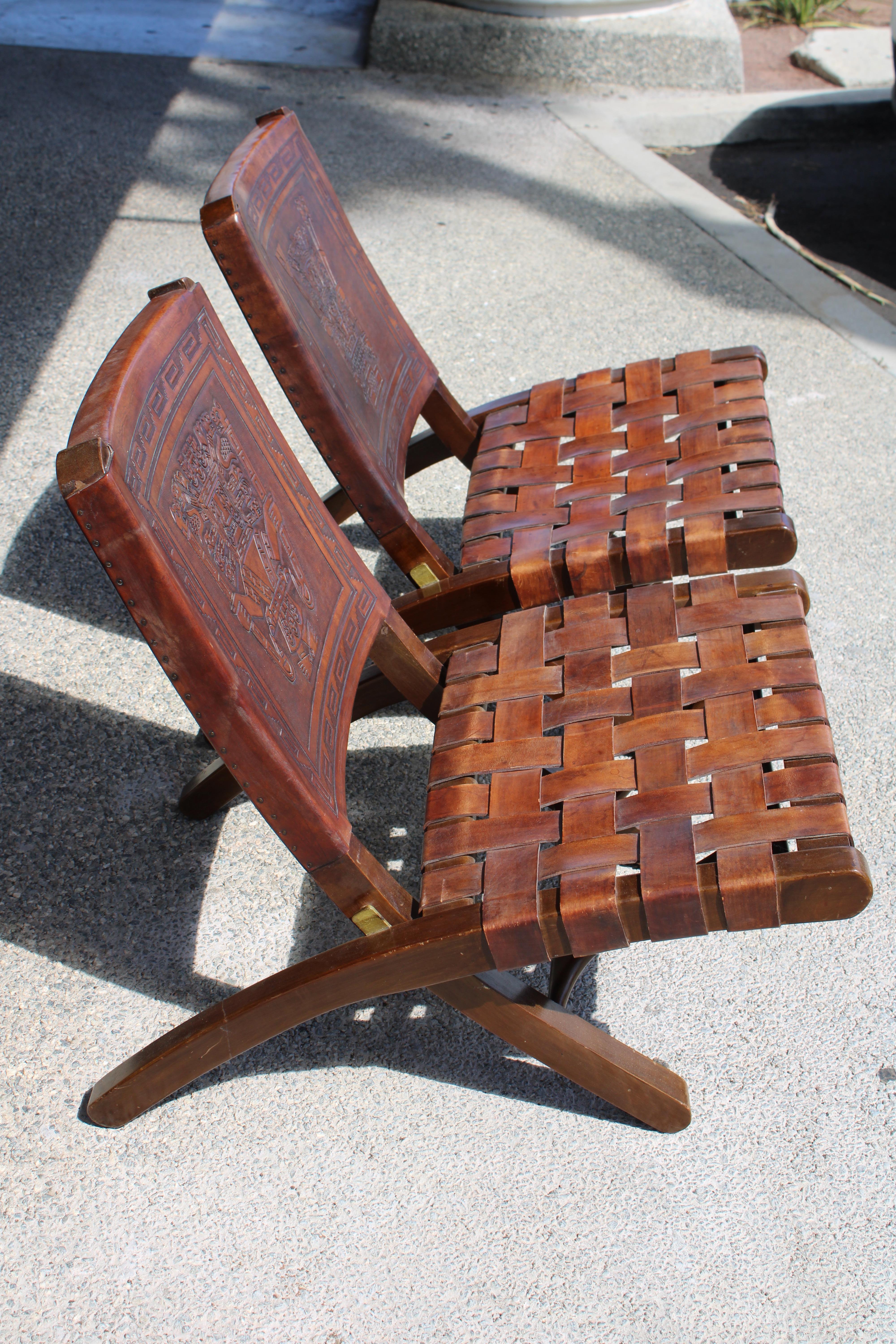 Pair of folding chairs by Angel Pazmino for Muebles De Estilo, Ecuador, circa 1960's. Wood frames with thick saddle leather backs embossed with Incan or Aztec designs. Seats are done in a woven piecrust pattern. Solid brass hinges. Chairs measure