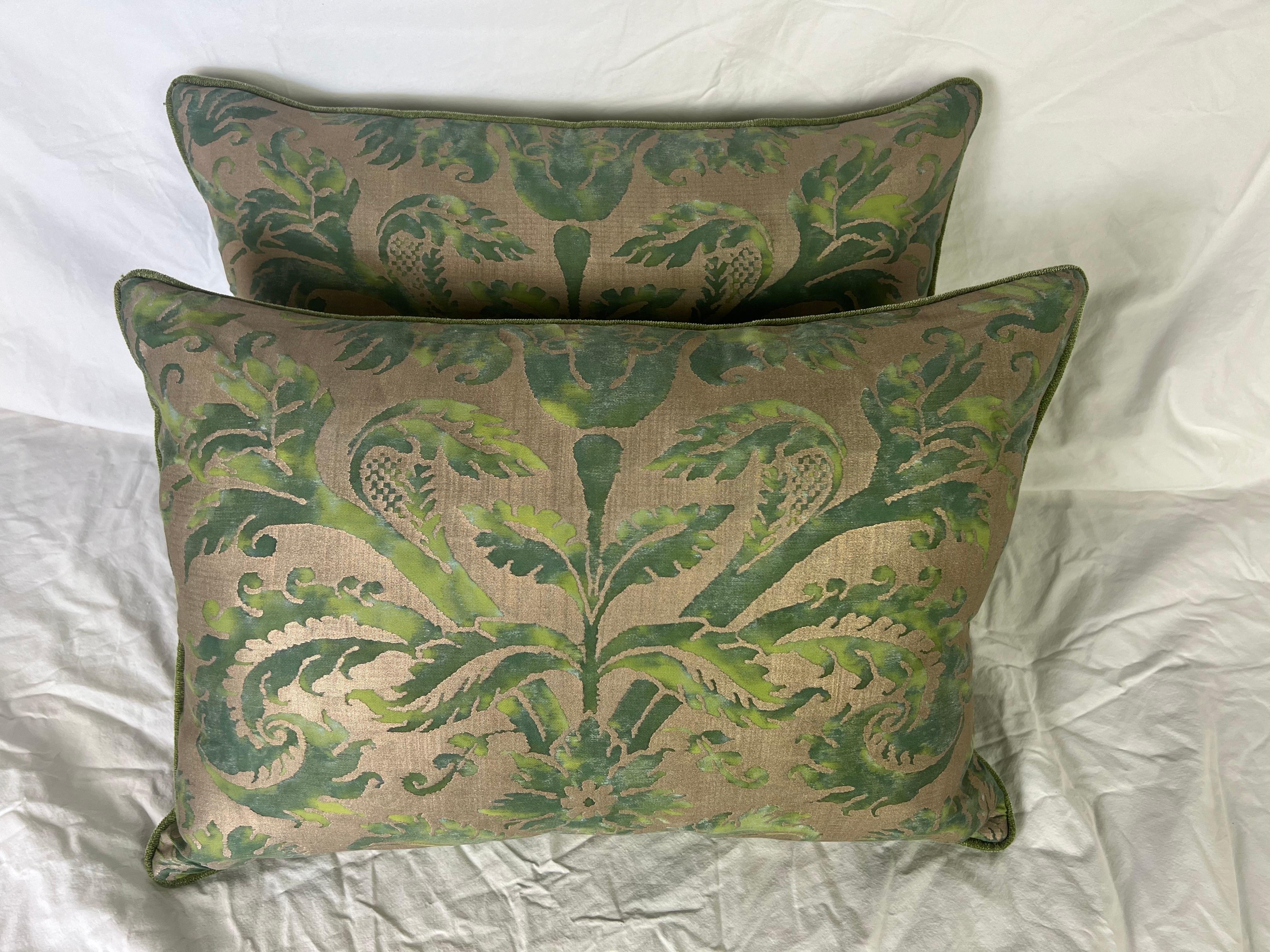These pillows are made from Fortuny fabric, which is known for its luxurious, hand-printed textiles. The design features an ornate, stylized botanical motif in various shades of green on a warm bronze-toned background. The pattern includes scrolling
