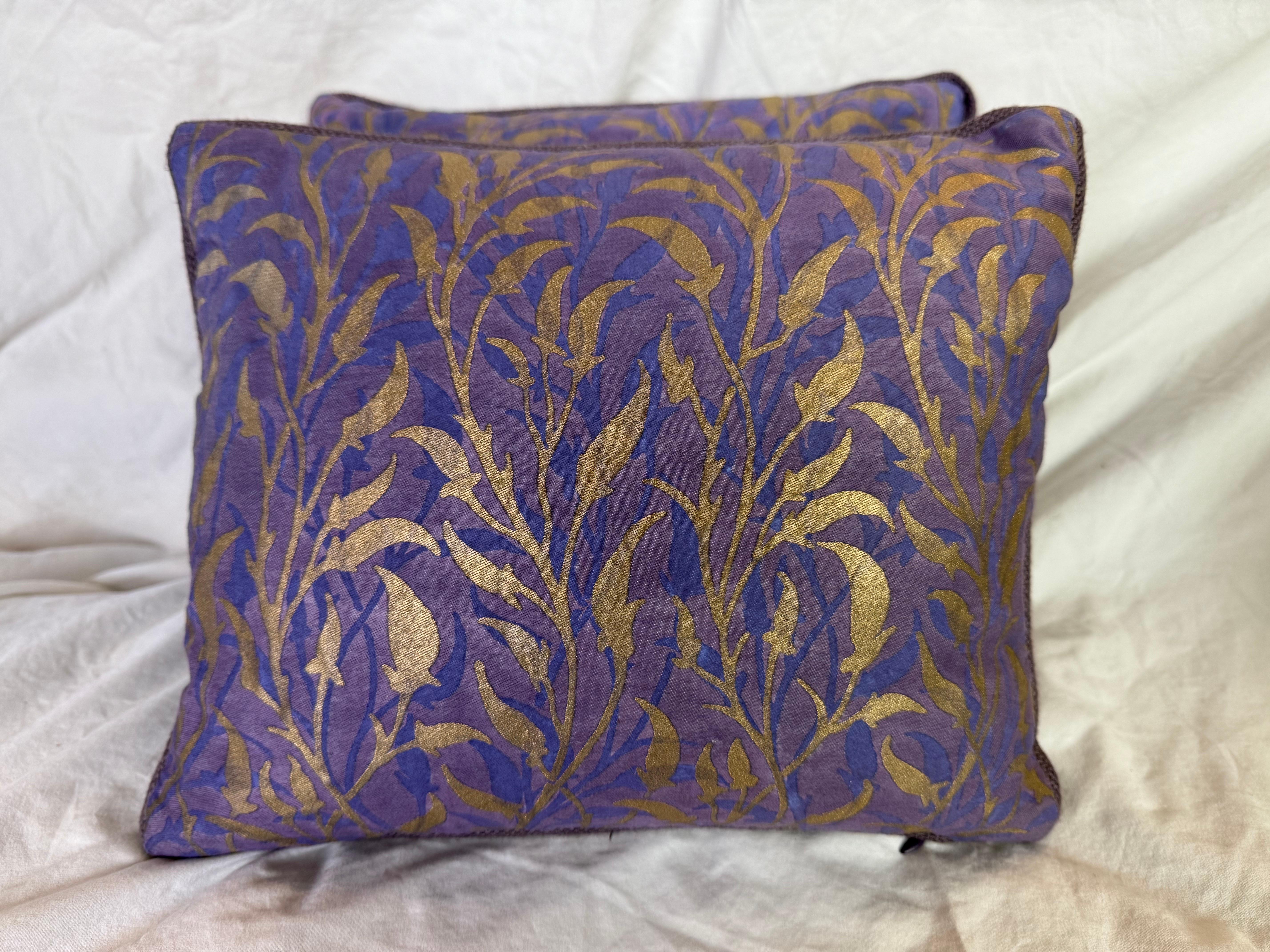 Estos cojines Fortuny presentan un diseño lujoso y elegante típico de la estética característica de la marca. El tejido presenta un atrevido motivo de hojas y enredaderas en un rico tono dorado, sobre un fondo morado intenso con matices azules. El