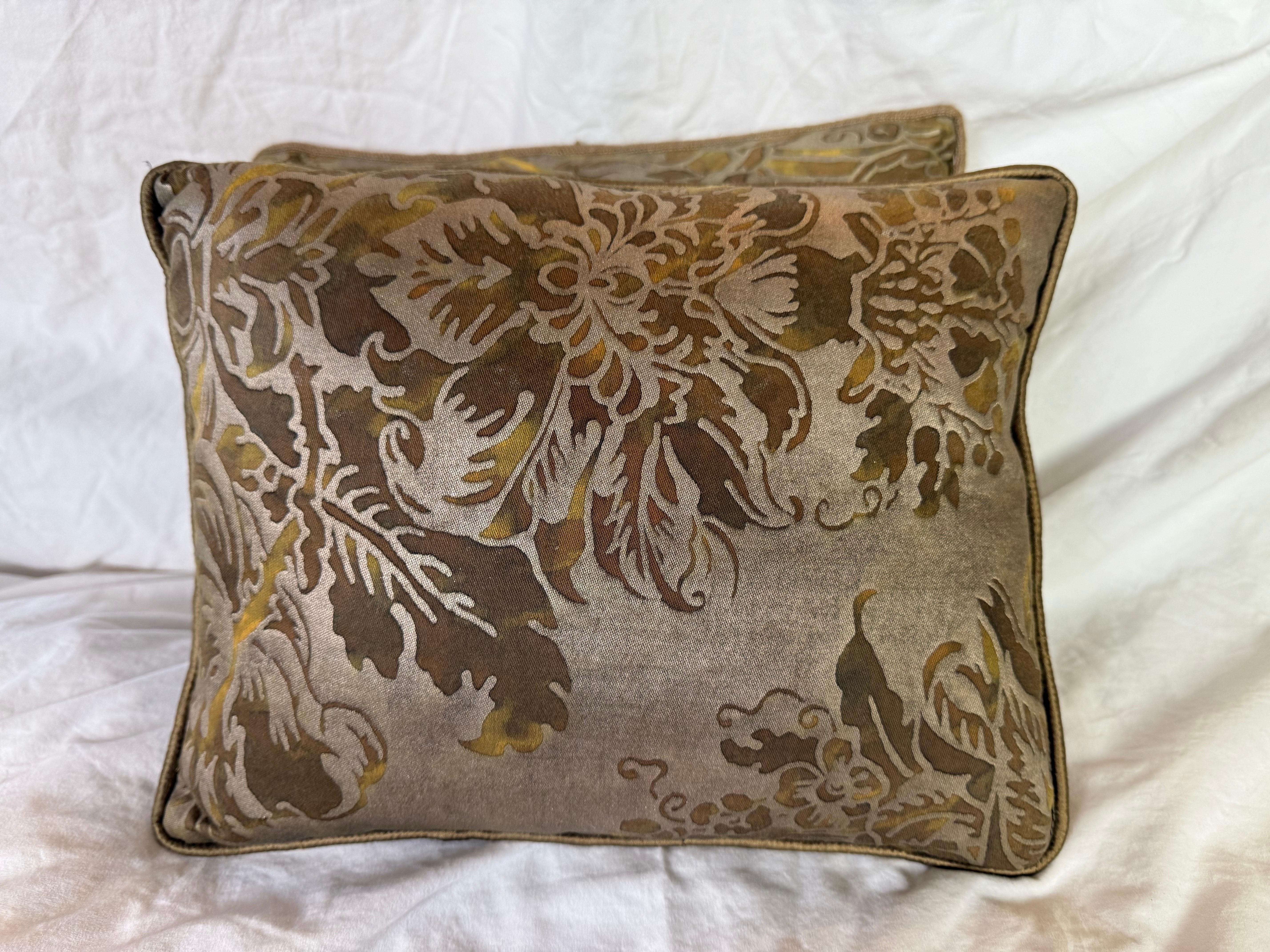 These Fortuny pillows feature an elegant botanical design in a soft, earthy palette. The fabric showcases a large-scale, floral and foliage motif in shades of gold, bronze, olive, and cream, printed against a taupe or warm gray background. The