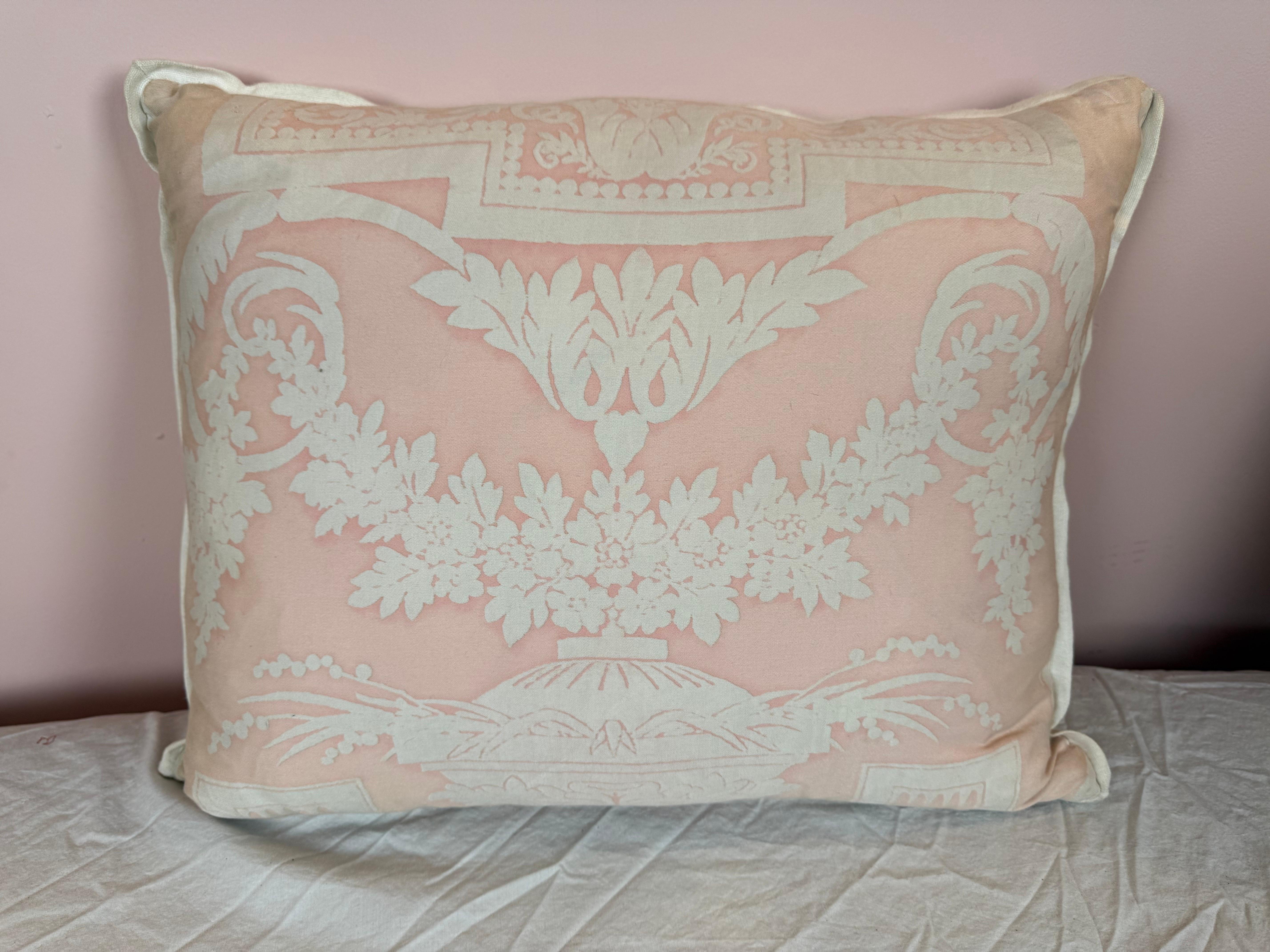 Questo cuscino presenta un design delicato ed elegante realizzato in tessuto Fortuny. Il tessuto presenta uno sfondo rosa tenue con un motivo classico bianco in stile damascato, caratterizzato da motivi floreali e ornamentali simmetrici che evocano