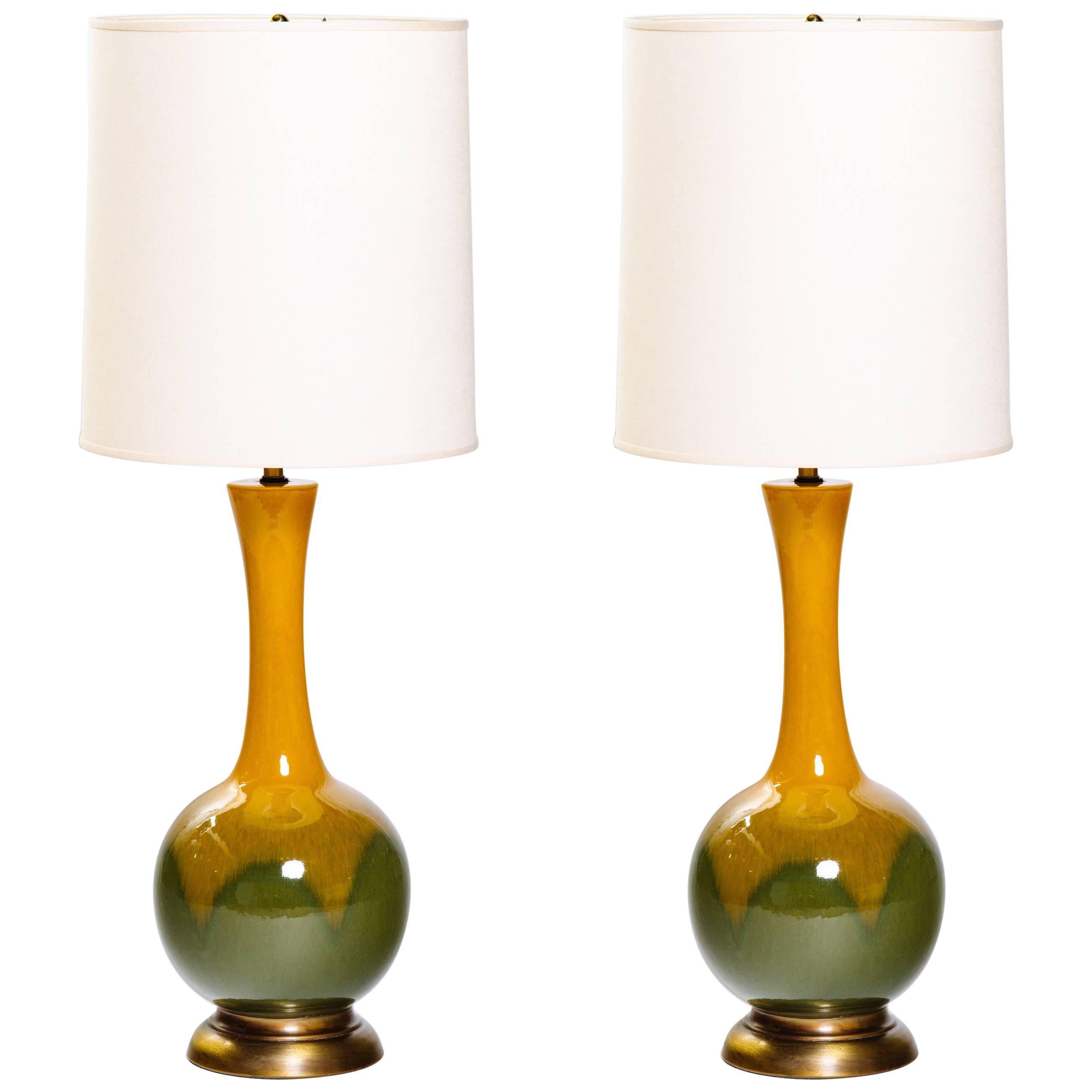 Gorgeous pair of tall ceramic pottery lamps with bulbous form and long neck design. The lamps have a glazed finish in gradient hues of mustard yellow and green moss. Lamps have dark brass base and fittings. Shown with custom tall drum shades in