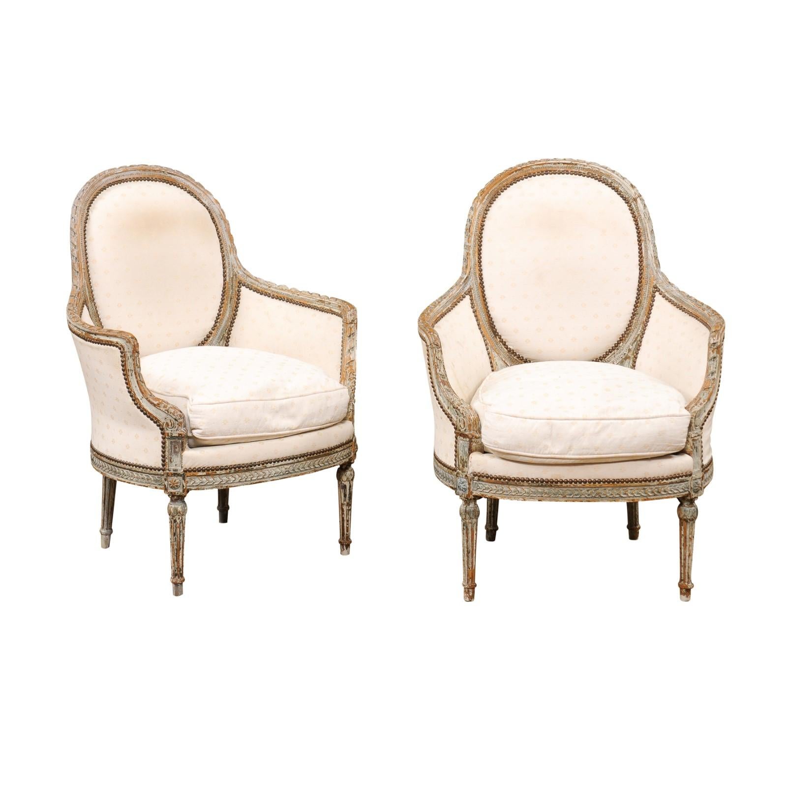 A pair of French Louis XVI style painted wood bergère chairs from the mid 19th century, with oval backs, carved twisted ribbon motifs, fluted legs, and distressed patina. Created in France at the beginning of Emperor Napoléon III's reign in the