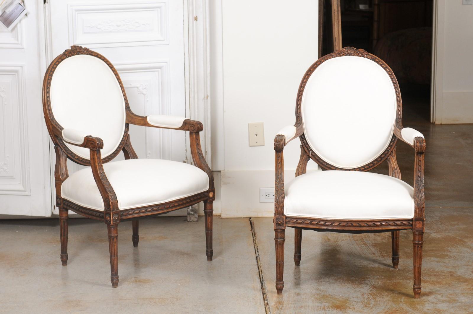 A pair of French Louis XVI style walnut armchairs from the mid-19th century, with oval backs, carved decor and new upholstery. Created in France during the reign of Emperor Napoleon III, this pair of armchairs presents the stylistic characteristics