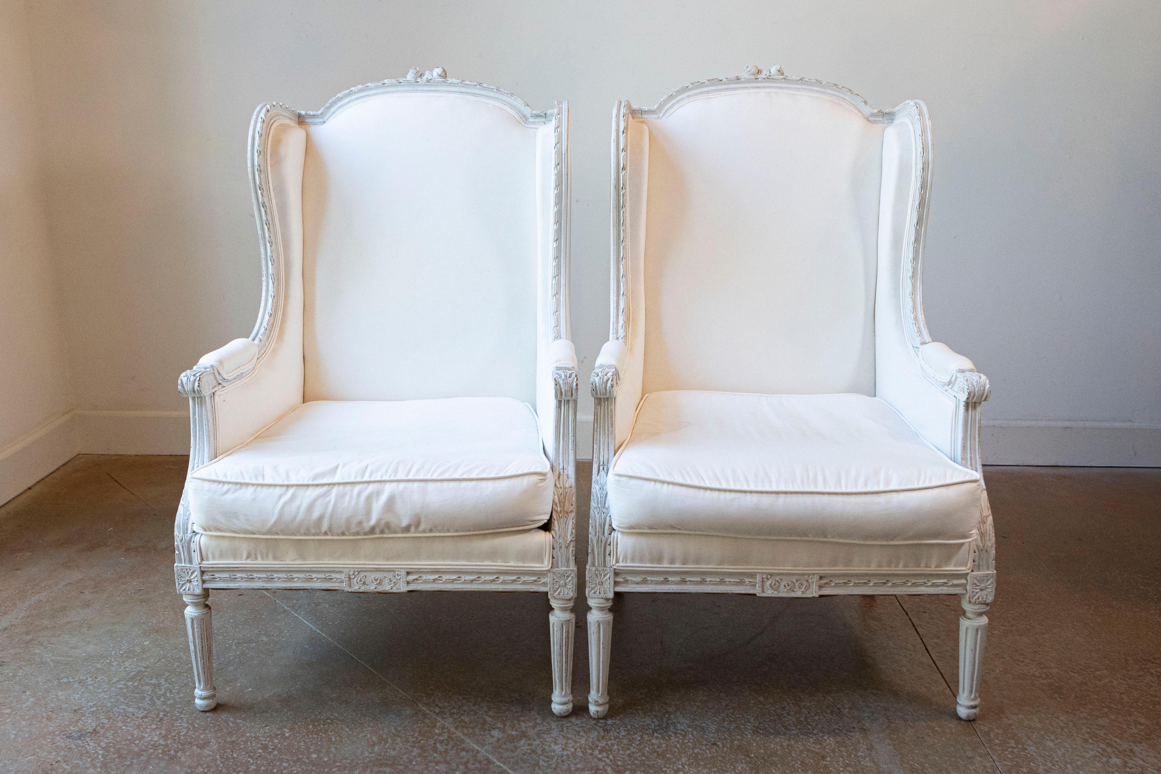 A pair of French Louis XVI style painted wood wingback bergère chairs from the late 19th century, with carved décor and new upholstery. Born in France during the later years of the 19th century, each of this pair of bergères features a winged back,