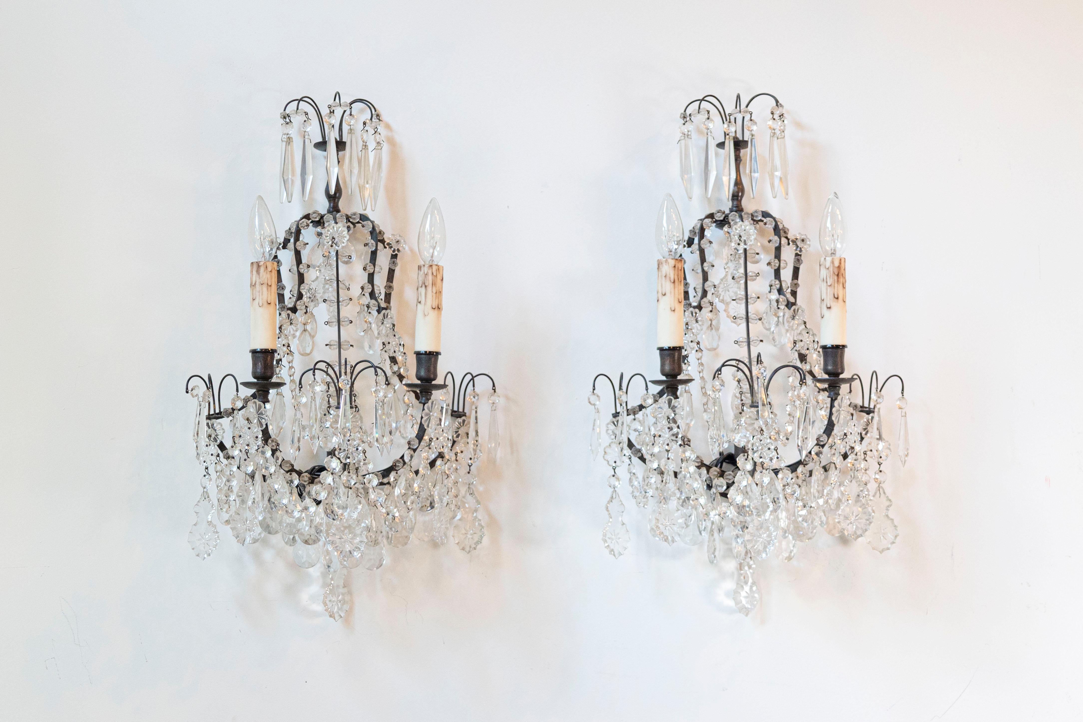 Paire d'appliques murales à deux lumières en cristal de style rococo français de la fin du XIXe siècle, avec des armatures en métal et des cristaux à facettes. Née dans les dernières années du XIXe siècle, cette paire d'appliques murales de style