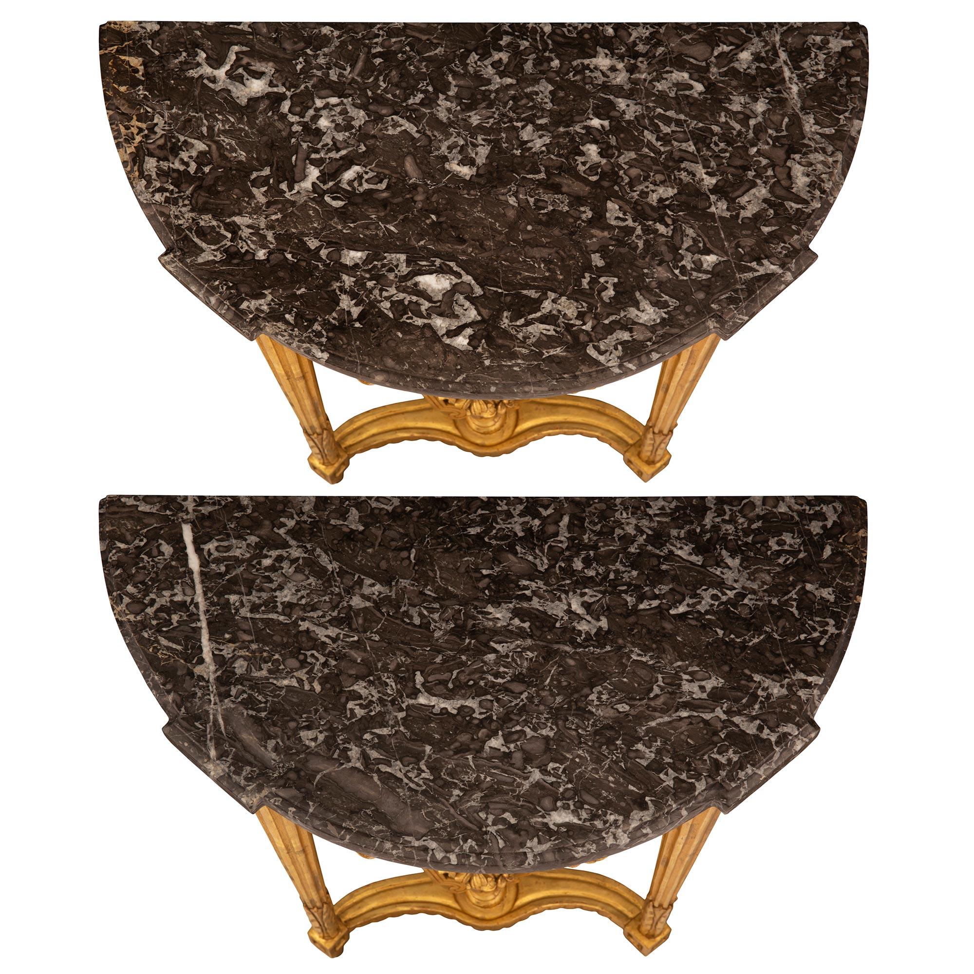 A most elegant and uniquely scaled pair of French 18th century Louis XVI period giltwood and Noir Antique marble consoles. Each small scaled wall mounted console is raised by circular tapered fluted legs with lovely foliate carvings and block