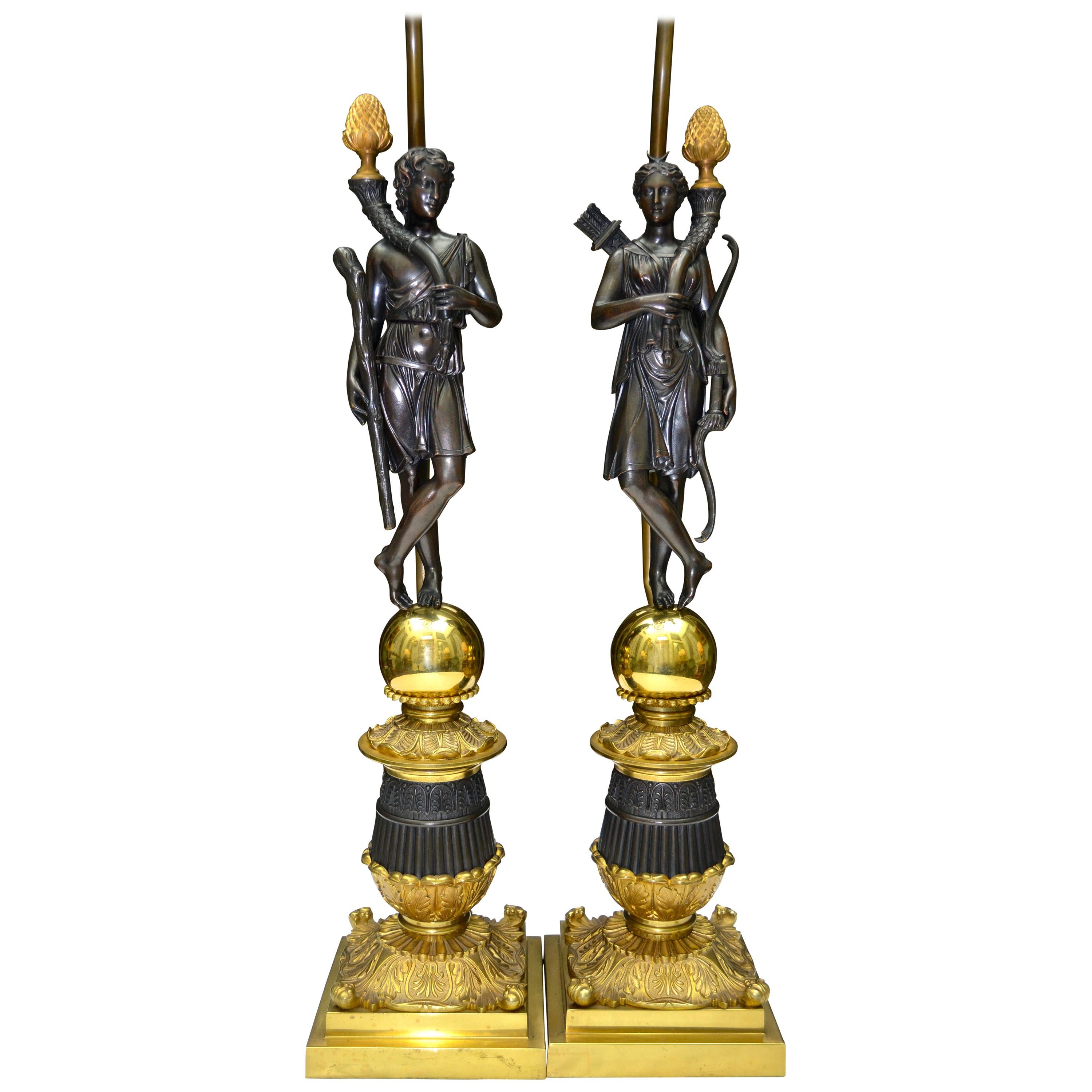 A pair of French restauration period figurative lamps bases featuring patinated bronze classically draped standing figures one representing Diana with her trademark bow and quiver of arrows, and the other a young Hercules with his club. Both are
