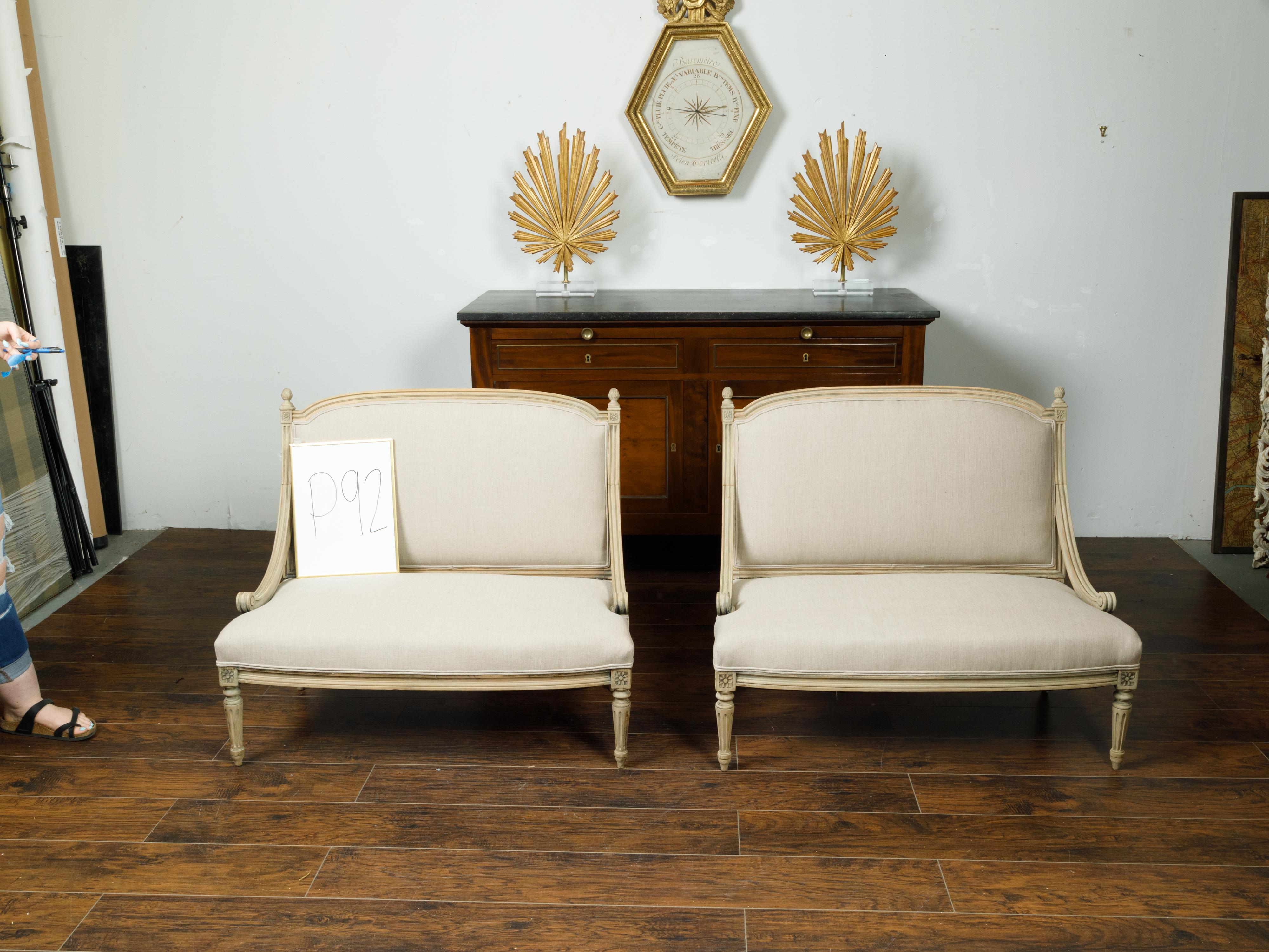 A pair of French Louis XVI style walnut settees from the early 20th century, with scrolling arm supports, fluted legs and new upholstery. Created in France during the early years of the 20th century, each of this pair of settees features a slightly