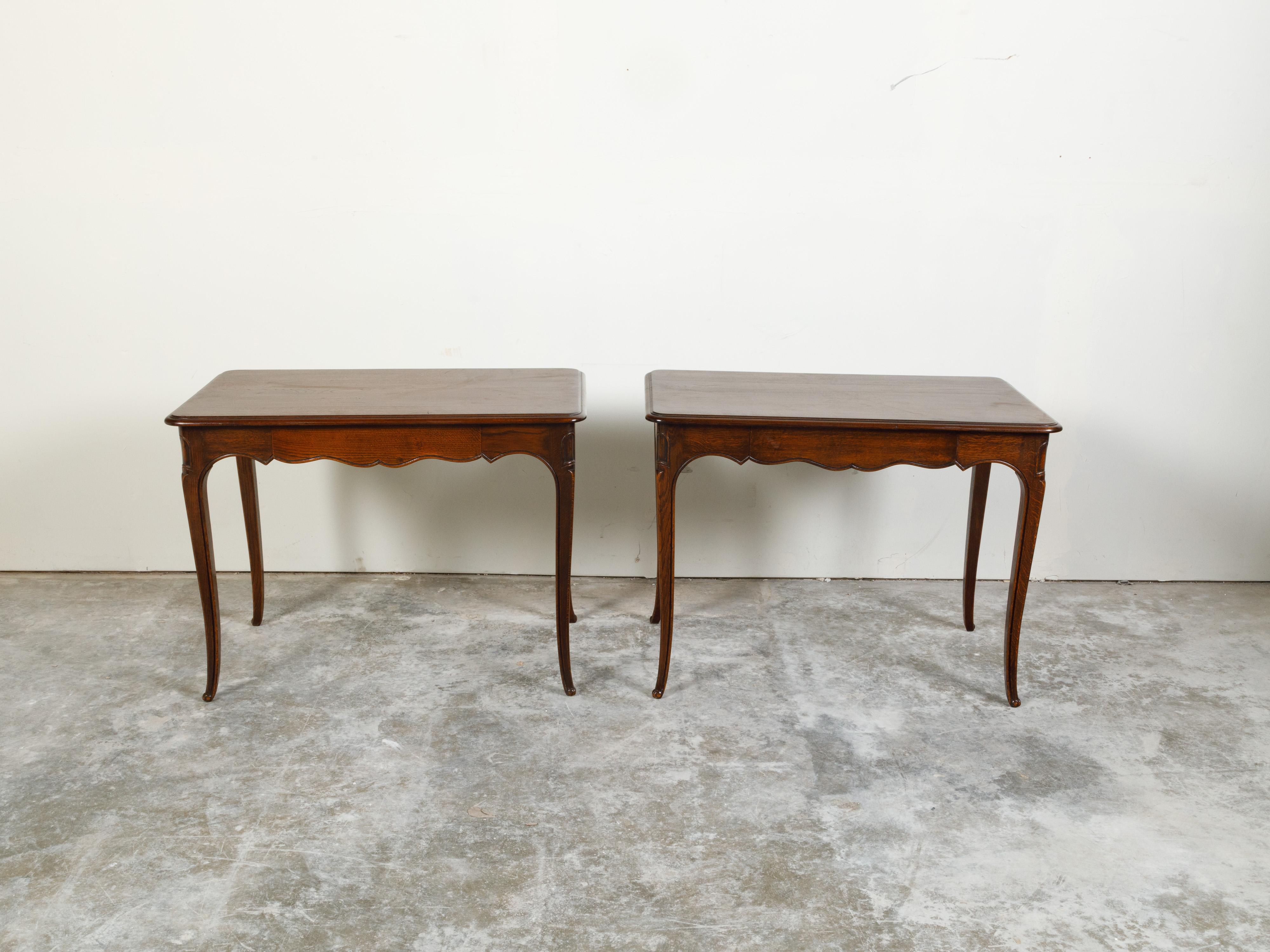 A pair of French oak console tables from the early 20th century, with single drawers, scalloped aprons and cabriole legs. Created in France during the Turn of the Century, each of this pair of console tables features a rectangular top with rounded