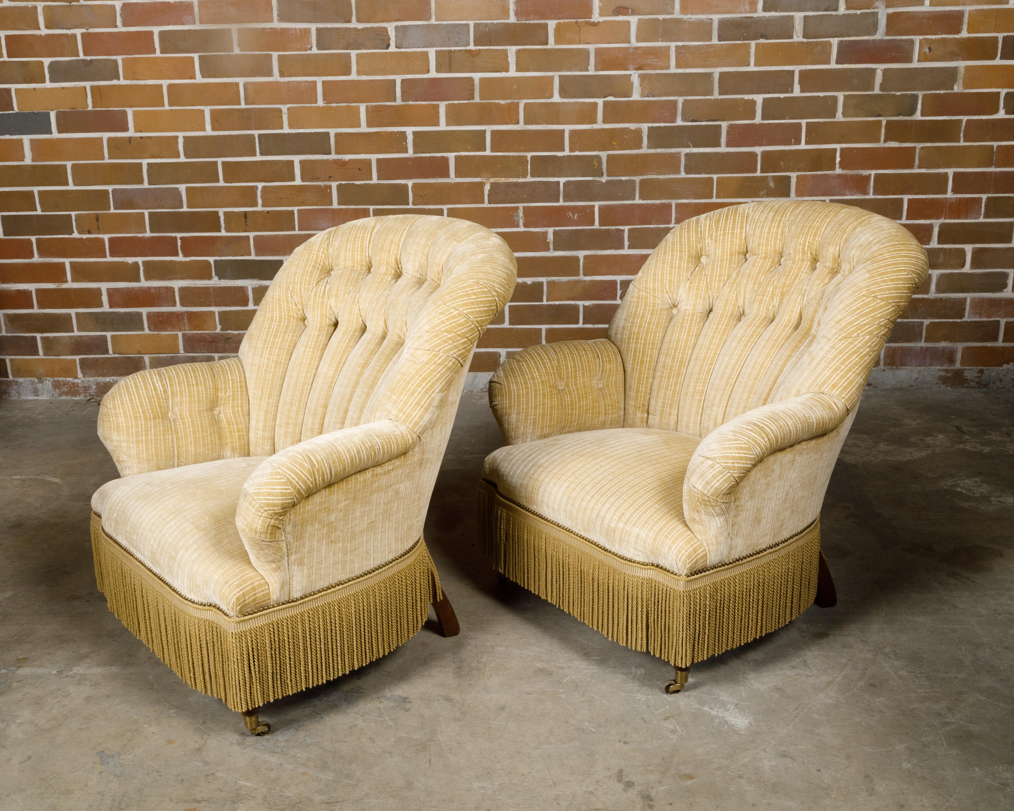 Paire de fauteuils crapauds français datant de 1920 environ, avec une tapisserie crème, des franges et des roulettes sur les pieds avant. Née en France vers 1920, cette exquise paire de fauteuils crapauds incarne l'élégance et le savoir-faire