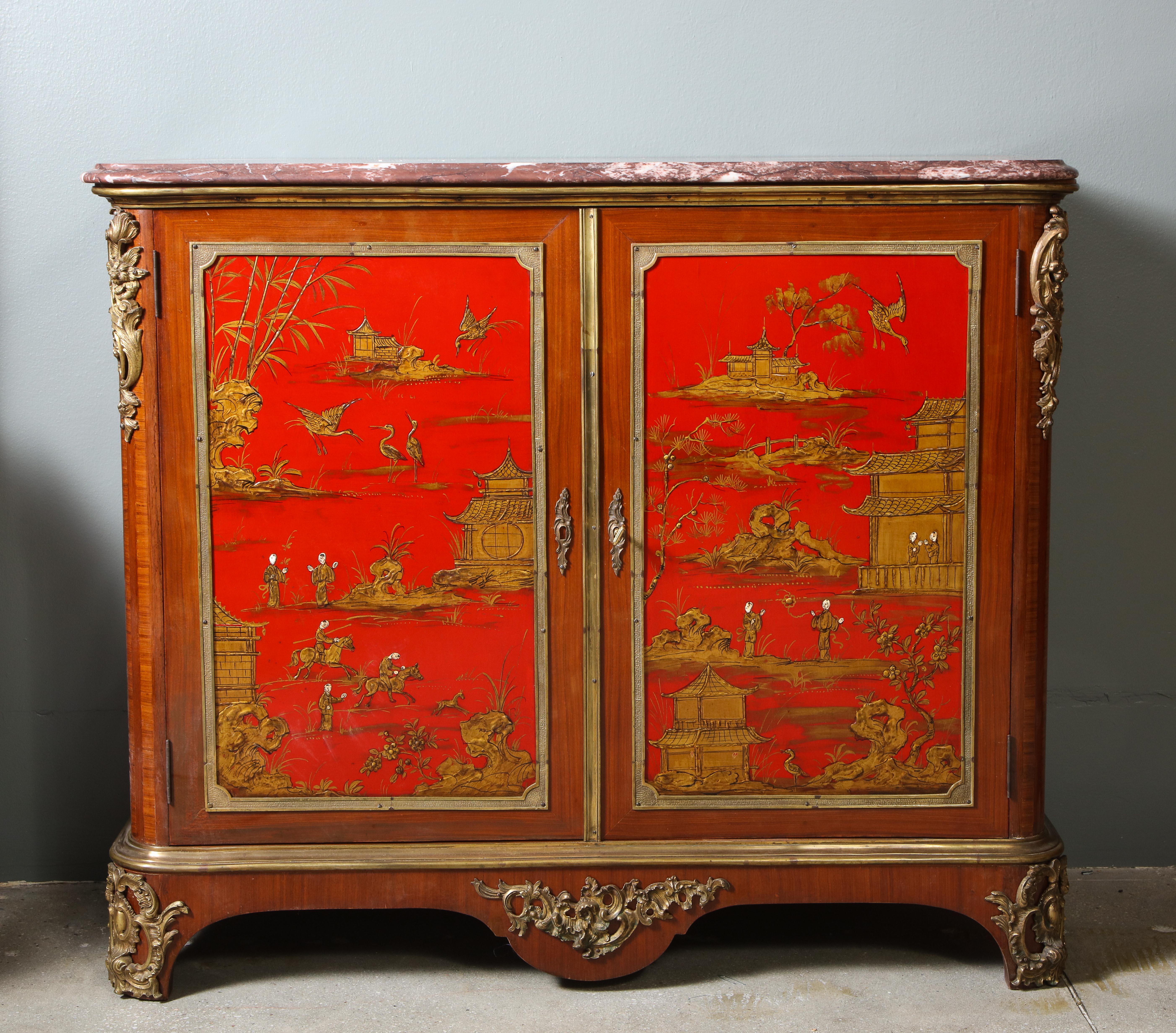 Each chinoiserie lacquered and gilt bronze mounted cabinet, attributed to Maison Jansen, with a marble top, over a pair of doors with bronze framed red lacquer panels, the sides with conforming decoration, the whole all over decorated with gilt
