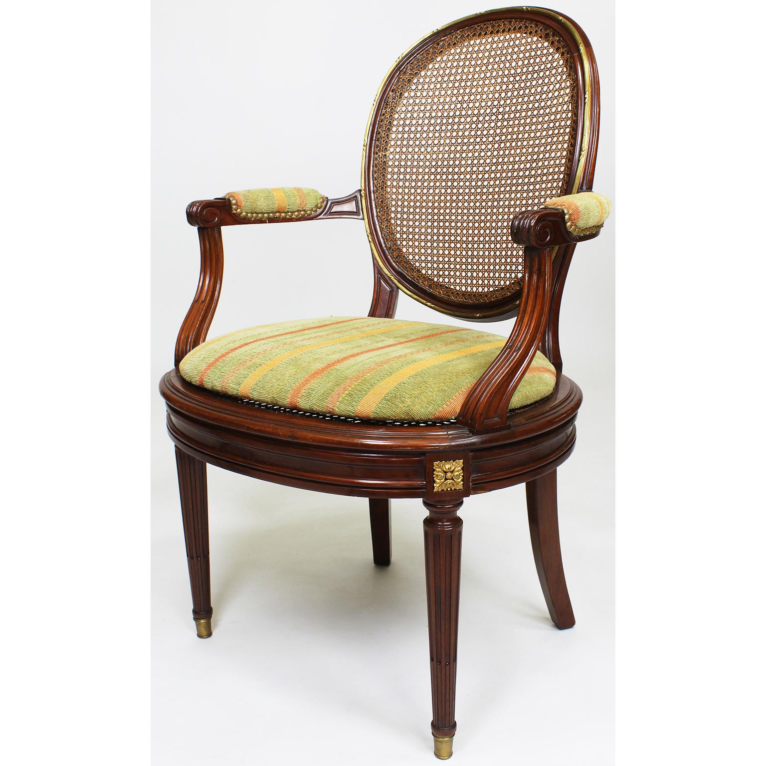 A fine pair of French 19th-20th century Louis XVI style Belle Époque mahogany and ormolu mounted dining or desk armchairs. The ovoid cane-backrests and seats, with padded open armrests, surmounted with a banded gilt-bronze trim and medallions,