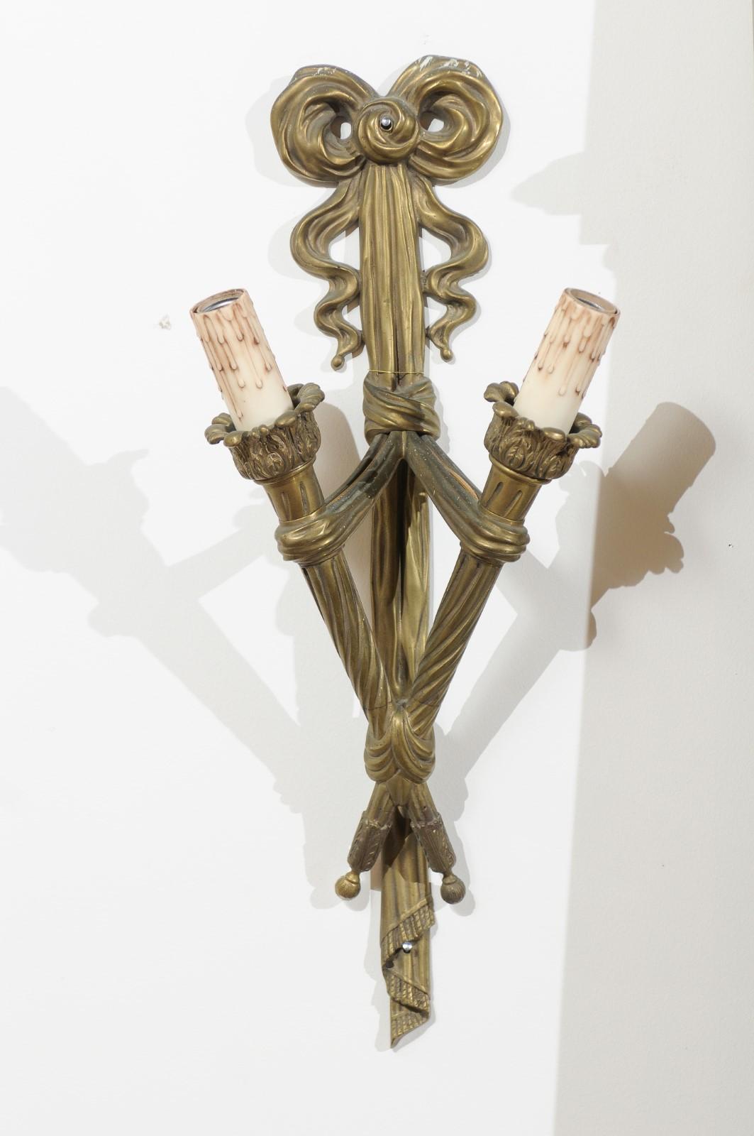 A pair of French two-light bronze sconces from the 19th century, with ribbon-tied torches. Born in France during the 19th century, each of this pair of wall sconces features two ribbon-tied torches supporting their candle sleeves and a foliage