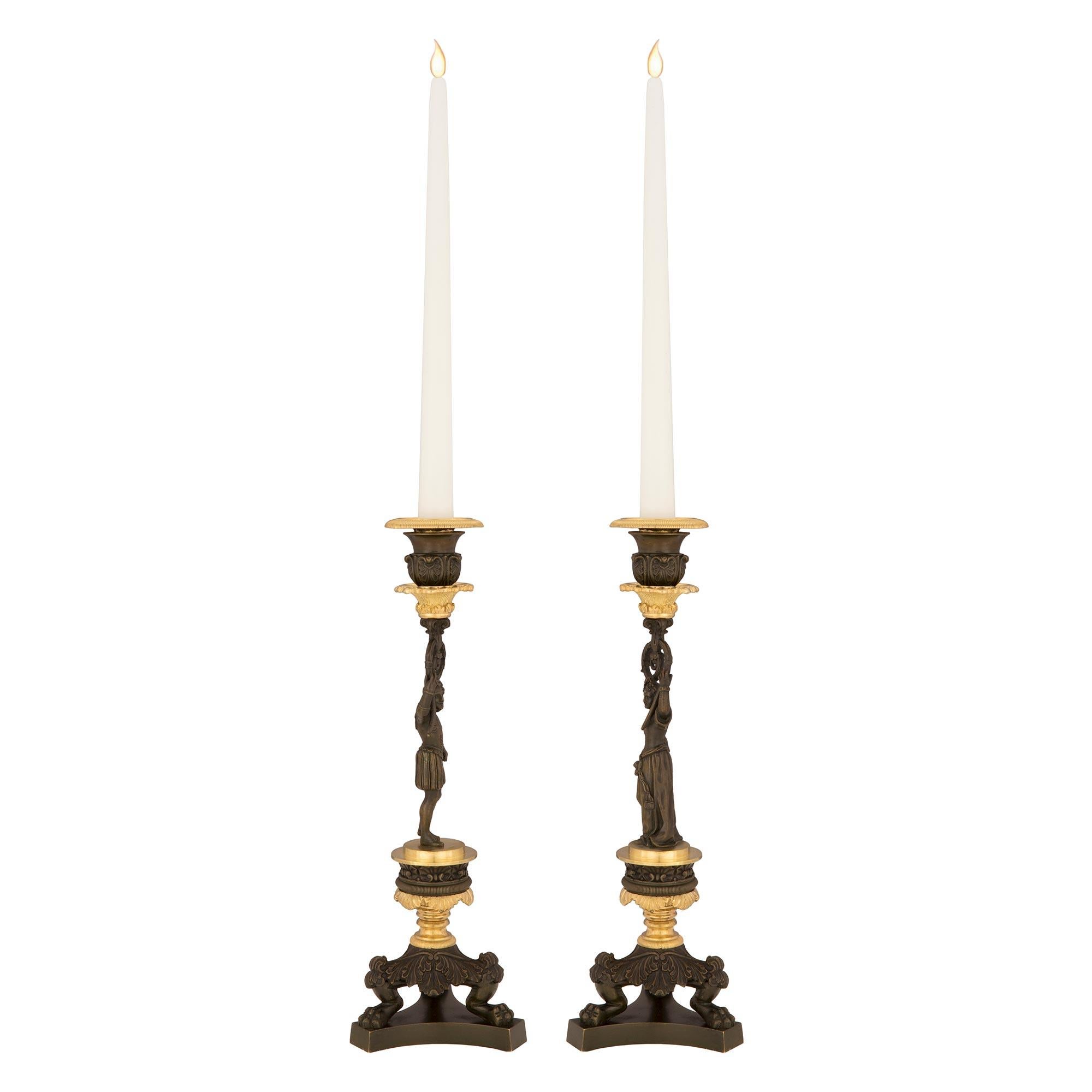 A striking pair of French 19th century Charles X st. patinated bronze and ormolu candlesticks. Each candlestick is raised by a triangular base with concave sides and handsome paw feet amidst acanthus leaves. Above, socle shaped supports and wrap