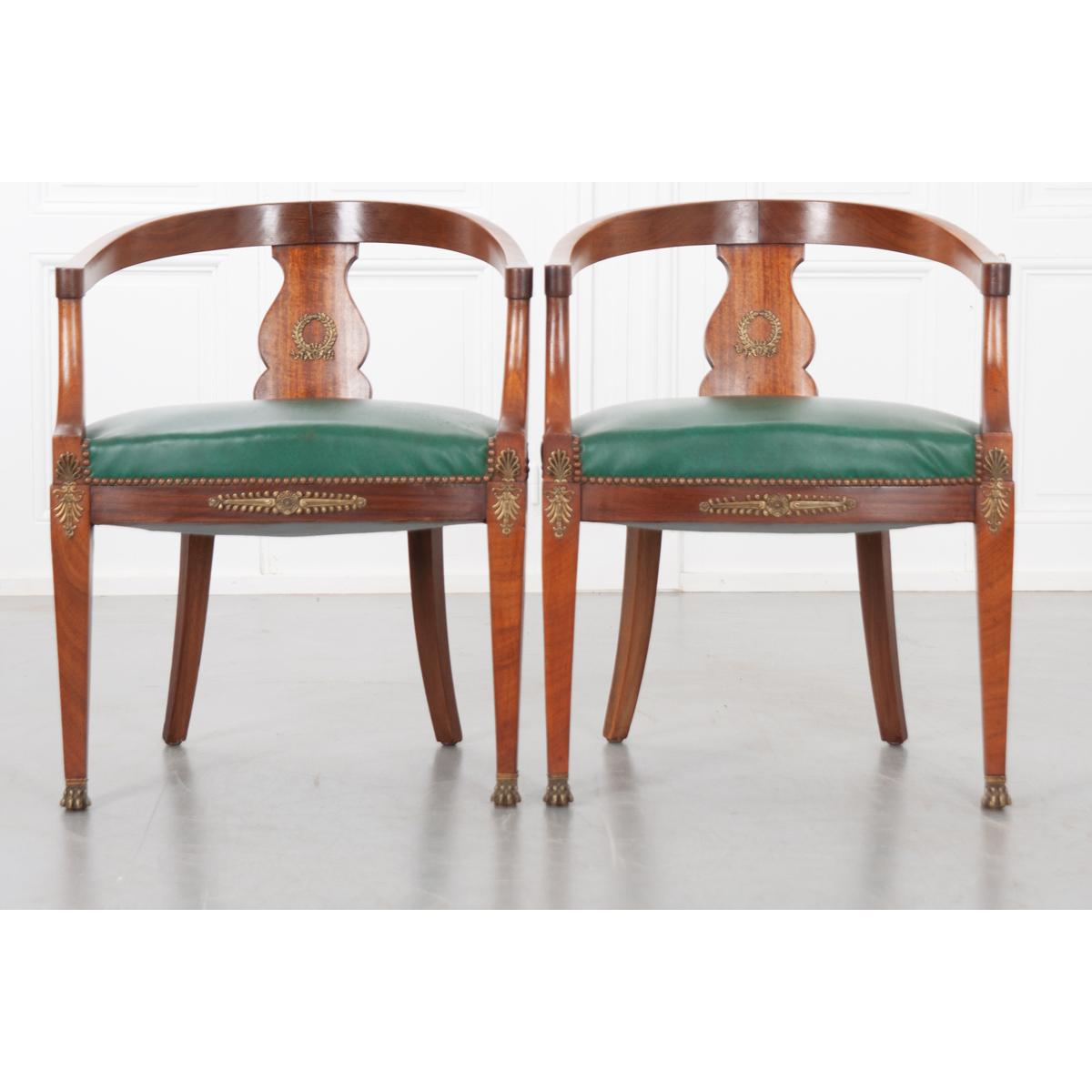 A superb pair of Empire-Style barrel back chairs. These classically styled mahogany chairs have green vintage vinyl upholstered cushions trimmed with a nailhead border. The concave back has a shaped backrest with an ormolu wreath at its center. The
