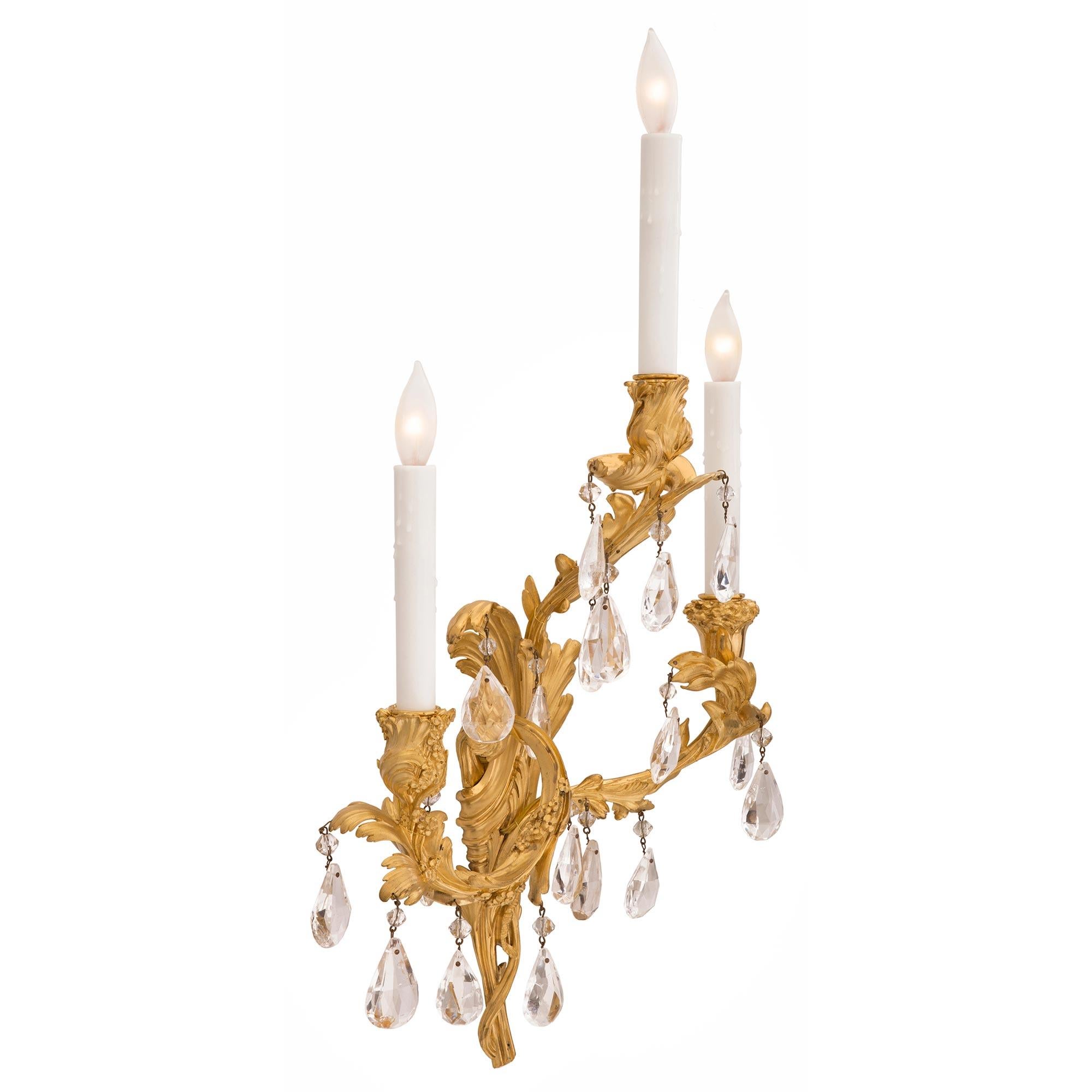A beautiful and very high-quality pair of French 19th century Louis XV st. Belle Époque period ormolu and rock crystal sconces. Each three-arm sconce is centered by a charming and most decorative pierced scrolled foliate design leading up the body
