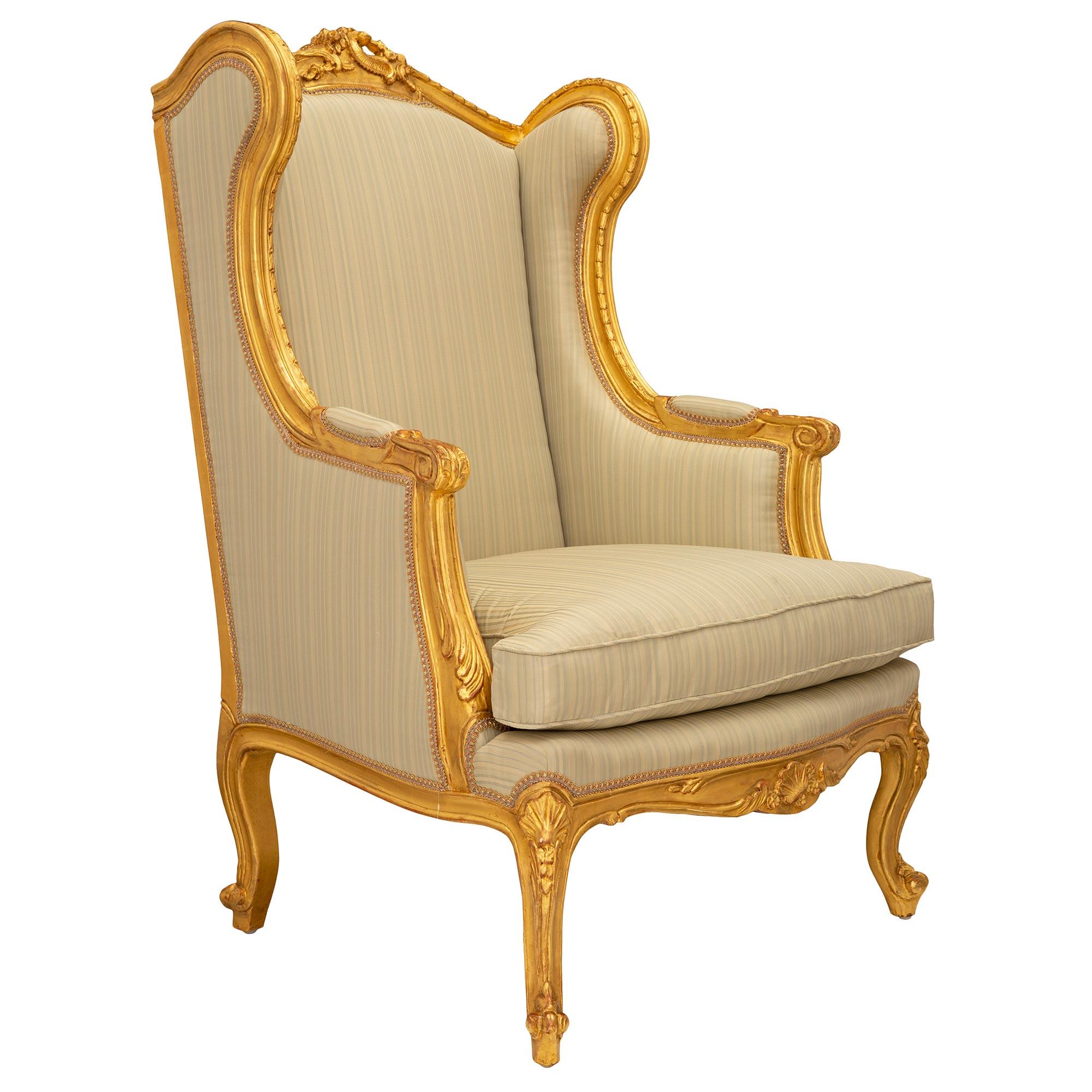 A beautiful pair of French 19th century Louis XV st. giltwood Bergères À Oreilles armchairs. Each armchair is raised by elegant cabriole legs with scrolled feet and finely carved seashell reserves at the corners. The arbalest shaped frieze displays
