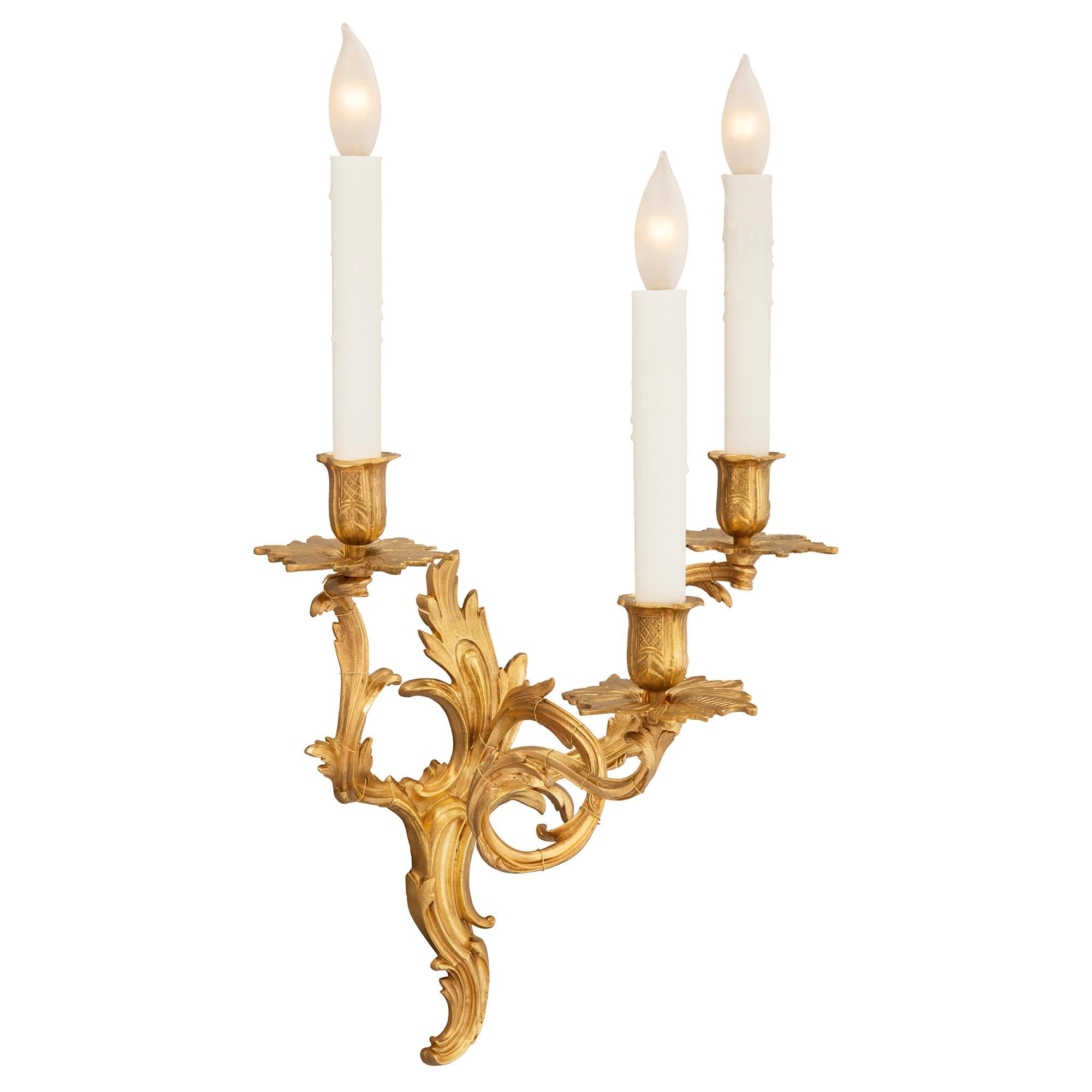 A charming true pair of French 19th century Louis XV st. ormolu three arm sconces. Each sconce is centered by a lovely scrolled foliate design in a striking satin and burnished finish which extends up the back plate. Branching out are the elegant