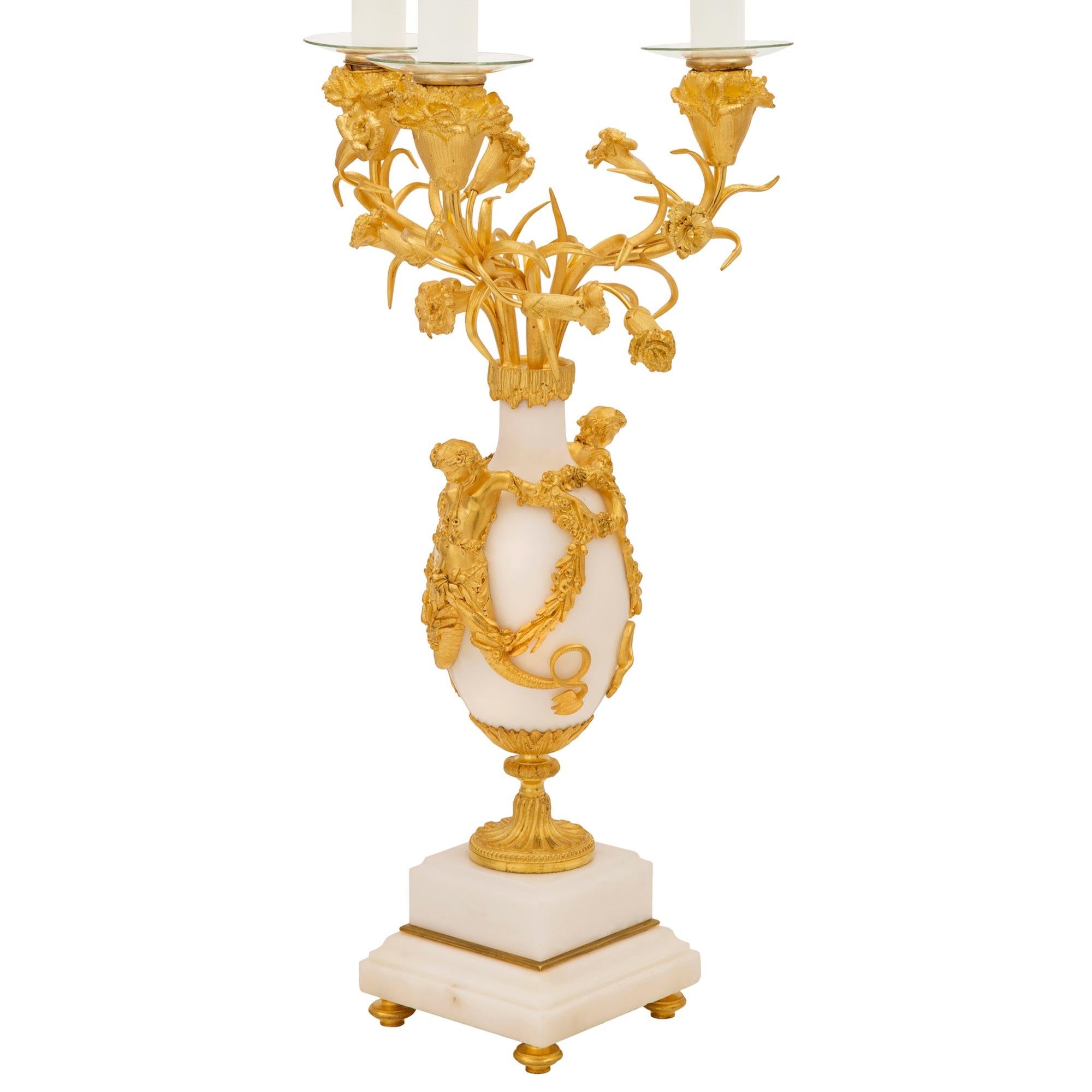 An exceptional pair of French 19th century Louis XVI st. Belle Époque period ormolu and white Carrara marble candelabra lamps. Each three arm lamp is raised by a square white Carrara marble base with an elegant stepped mottled design and fine topie
