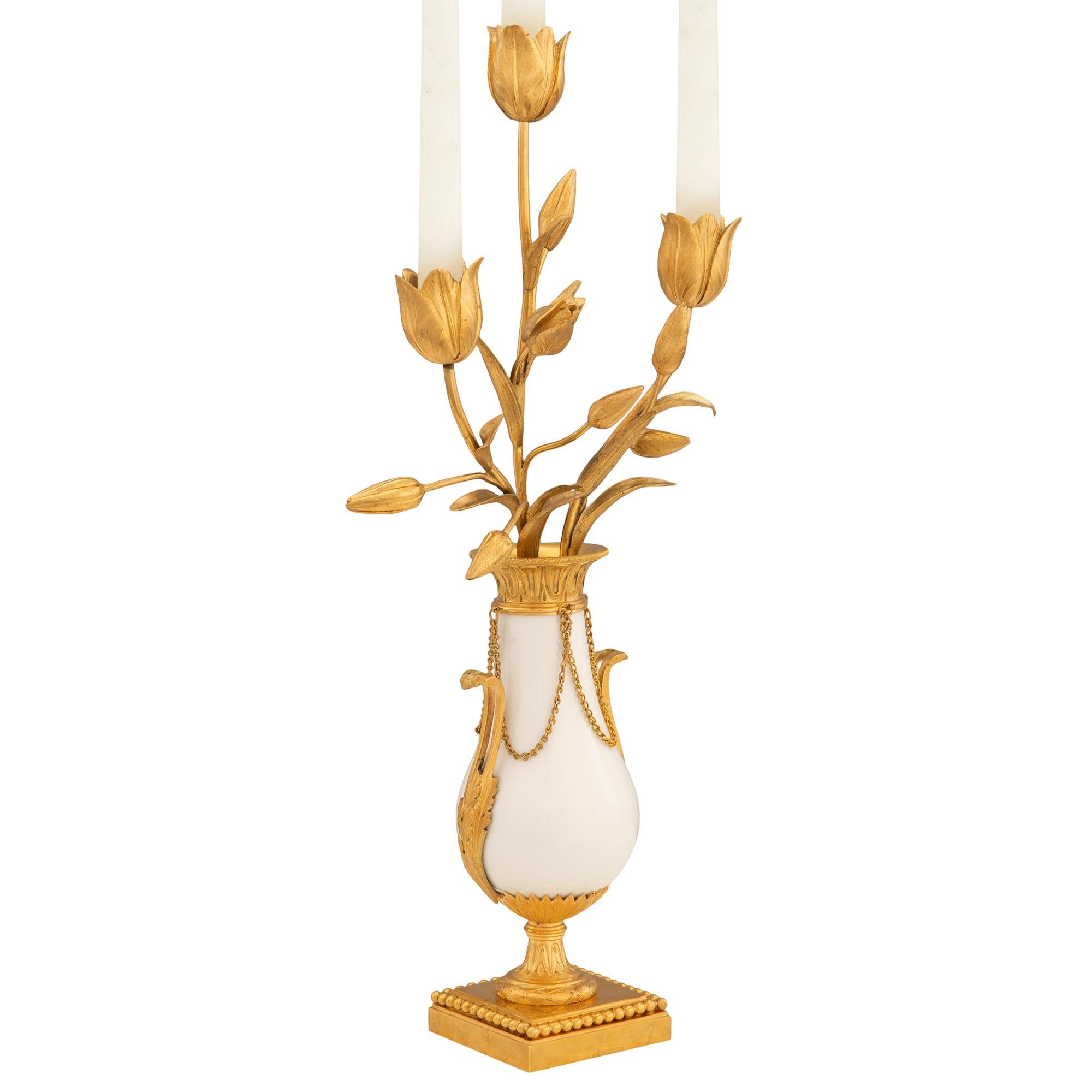 Un'elegantissima coppia di candelabri francesi del XIX secolo in stile Luigi XVI in ormolu e marmo bianco di Carrara. Ogni candelabro a tre bracci è sollevato da una base quadrata in ormolu con una raffinata fascia di perline avvolgente sotto il