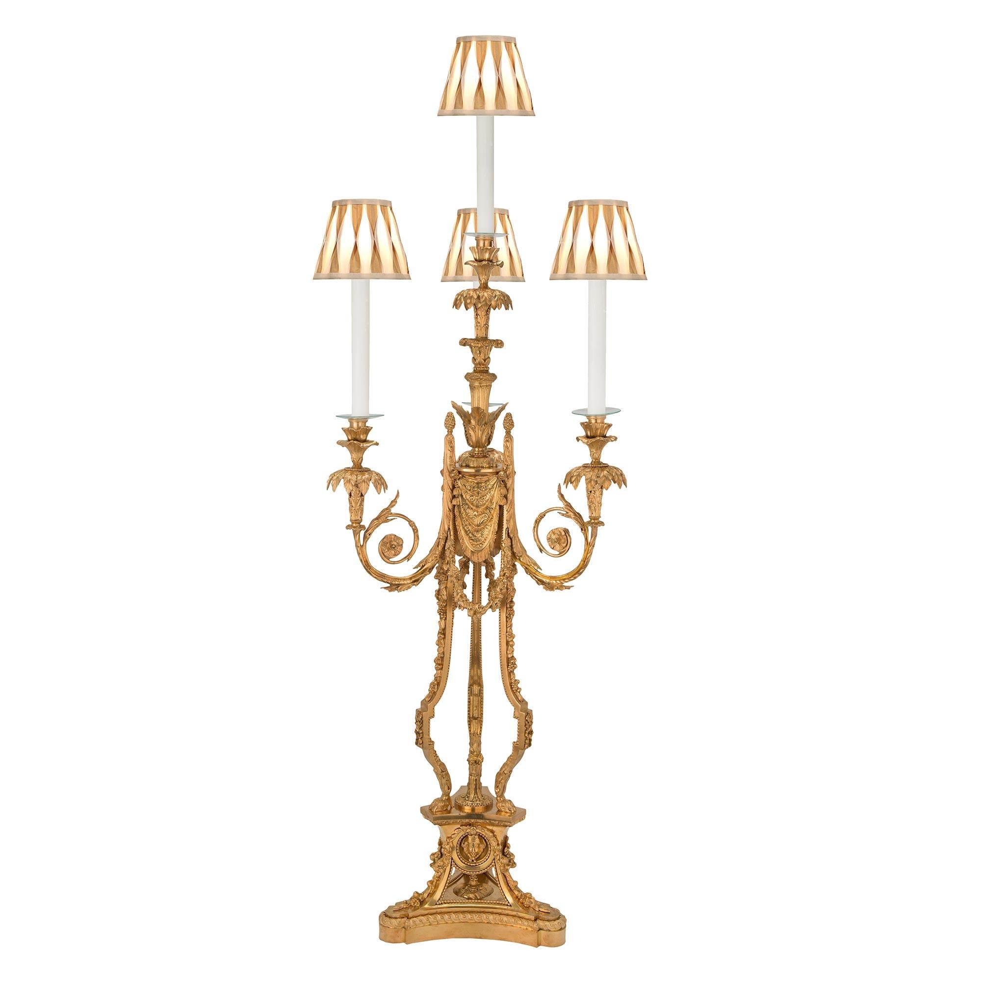 A stunning and very large scale pair of French early 19th century Louis XVI st. ormolu candelabras mounted into lamps after a model by Pierre Gouthière. Each lamp is raised by a triangular shaped base with rounded corners and concave sides. The base