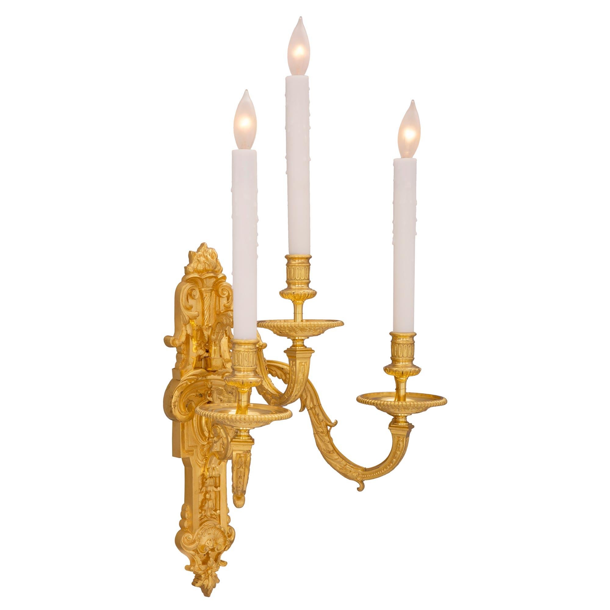 A striking pair of French 19th century Louis XVI st. ormolu sconces. Each three arm sconce is centered by a richly chased and wonderfully detailed backplate with a bottom seashell reserve, lovely foliate designs and an impressive eternal flame above