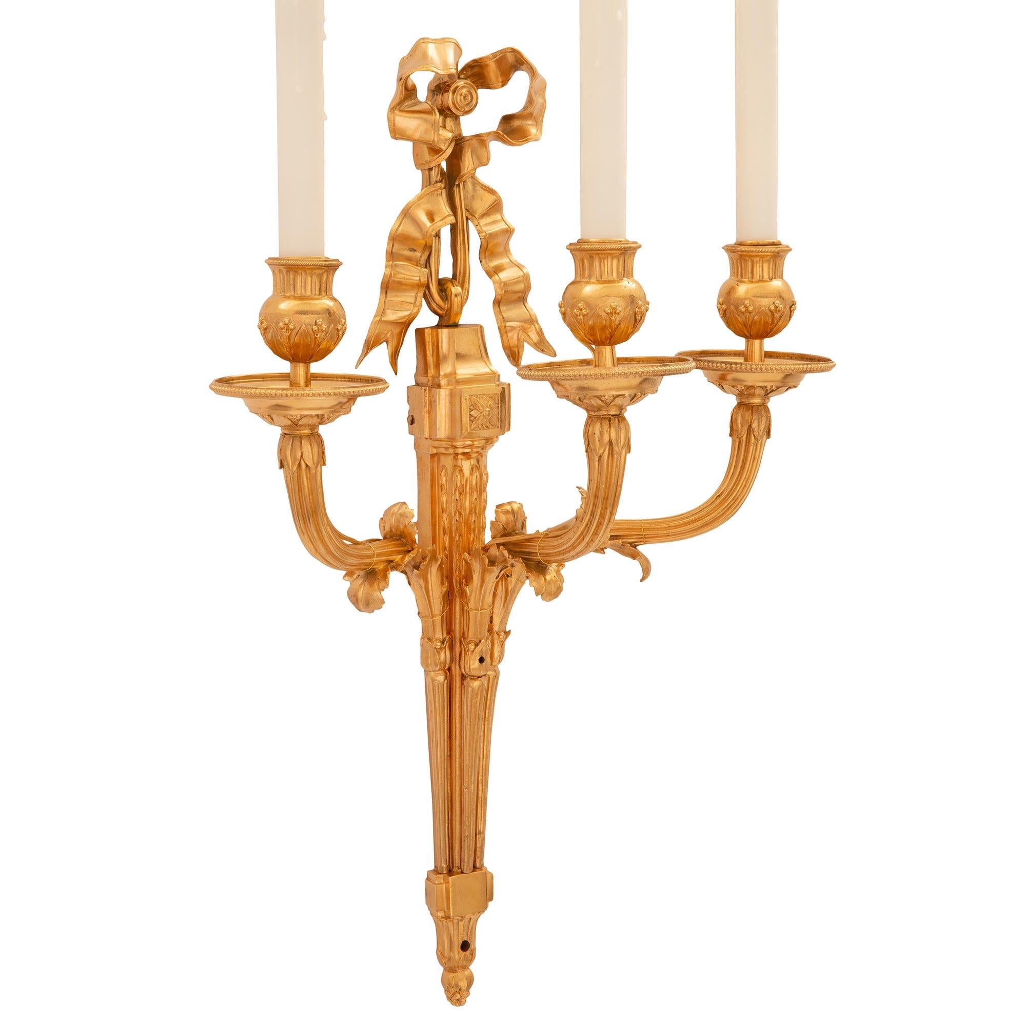 An extremely elegant pair of French 19th century Louis XVI st. ormolu sconces. Each three arm sconce is centered by a finely detailed bottom acorn finial below the elegant tapered fluted body and striking block rosette from where a charming and