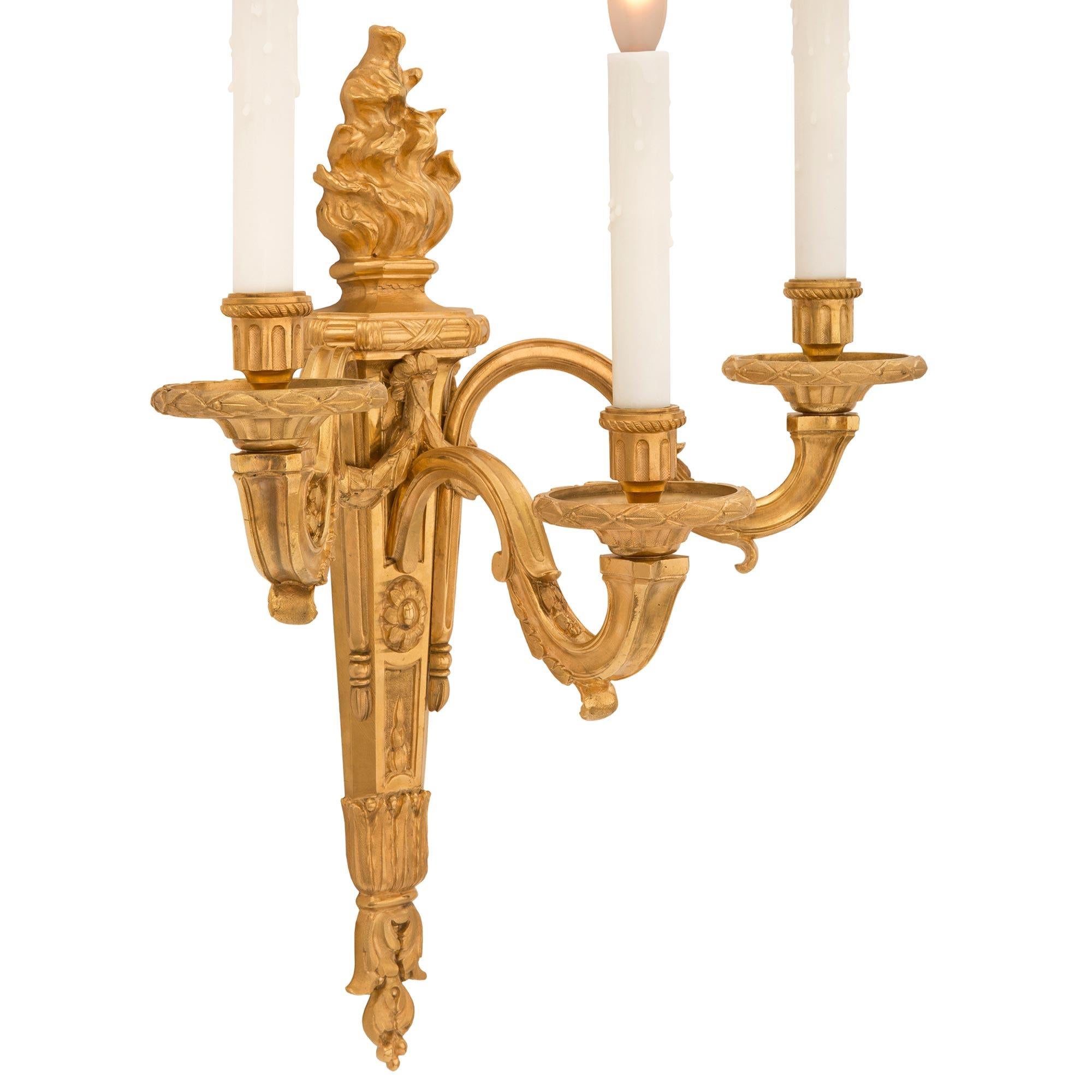 A striking pair of French 19th century Louis XVI st. ormolu sconces. Each three arm sconce is centered by a charming bottom foliate finial below the elegant tapered body decorated with finely detailed foliate designs, a charming floral reserve,