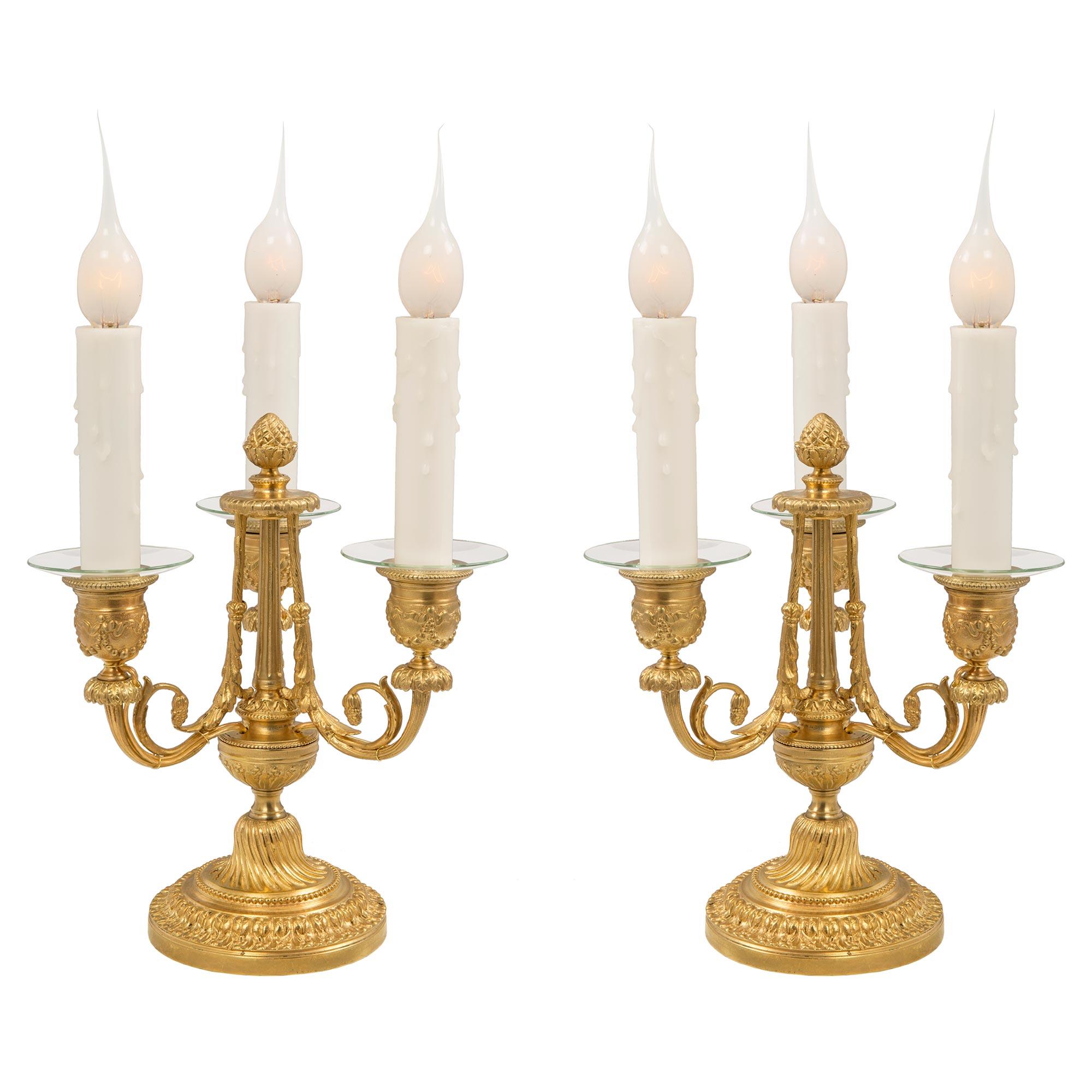 Une élégante paire de lampes candélabres à trois bras en bronze doré de style Louis XVI du 19ème siècle. Chaque lampe est surélevée par une base circulaire à gradins avec une belle bande de feuillage enveloppante et un motif perlé. Au-dessus du