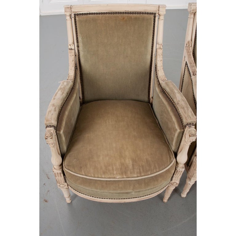 A fabulous pair of hand-carved, painted bergères. This pair features spectacular carved details that grace many parts of the cream-colored painted frame. These chairs are upholstered in a light green, worn fabric secured to the frame with brass