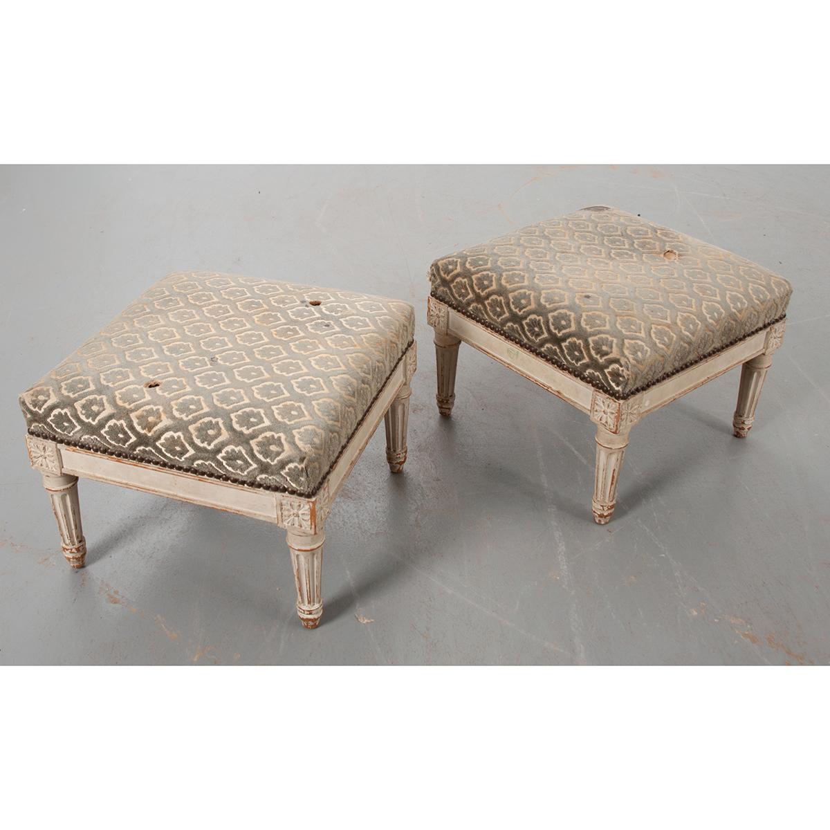 A pair of French 19th century Louis XVI-style foot stools. The worn upholstery is secured on the stool with a nailhead trim. They have their original painted finish. Rosettes are carved on each corner of the foot stools. All sit on turned fluted