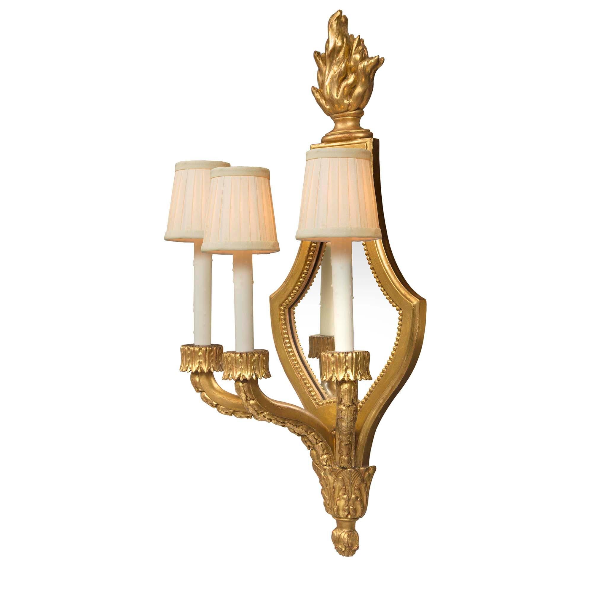 An elegant pair of French 19th Century Louis XVI st. giltwood three-arm mirrored sconces. Each sconce is centered by a fine bottom acorn inverted finial below large acanthus leaves. The three S-scrolled arms display finely carved berried laurel