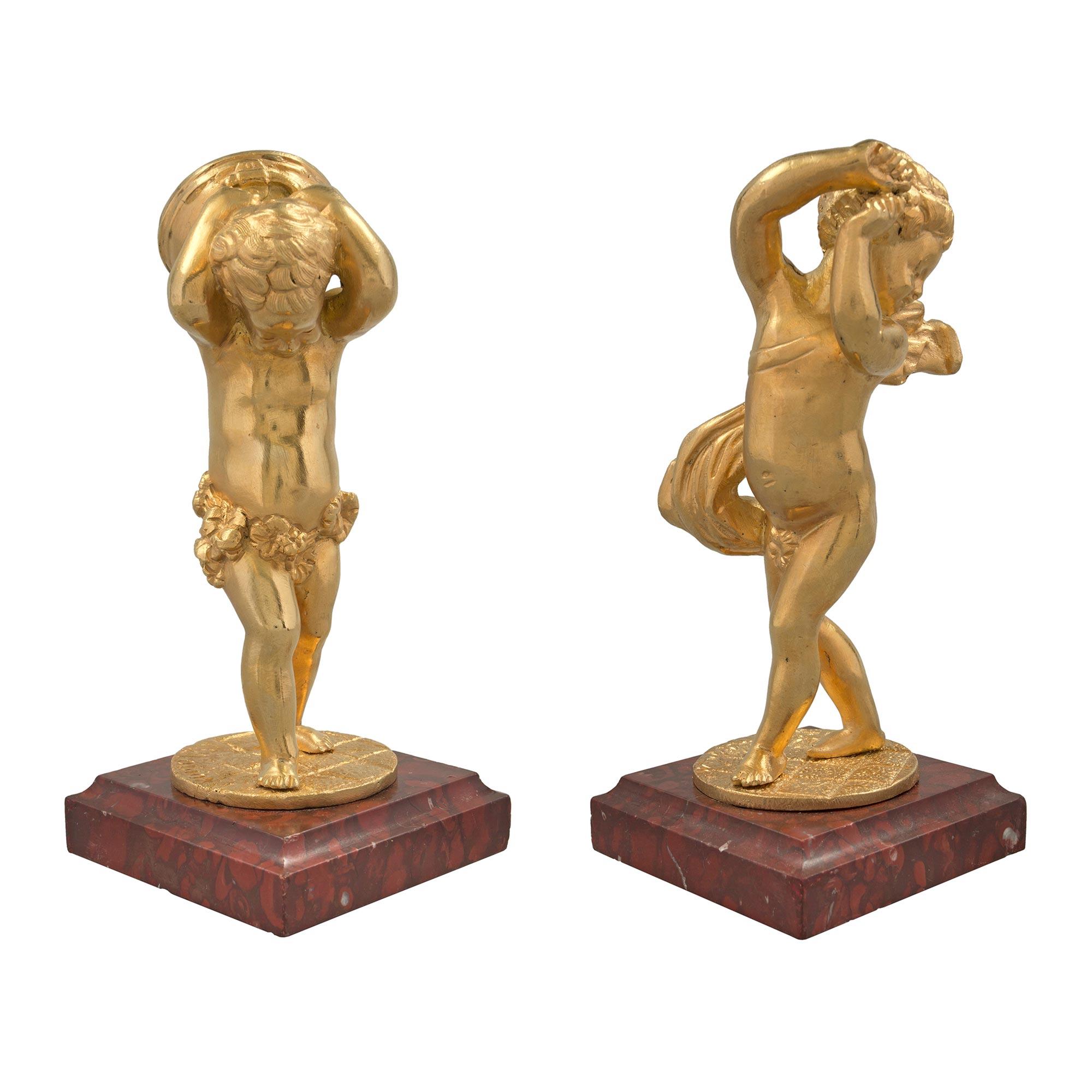 A charming pair of French 19th century Louis XVI st. ormolu cherub signed statues. The pair are raised by ormolu bun feet below a square Rouge Griotte marble base with mottled border. Above are the sweet ormolu cherubs, one lifting a drum behind his