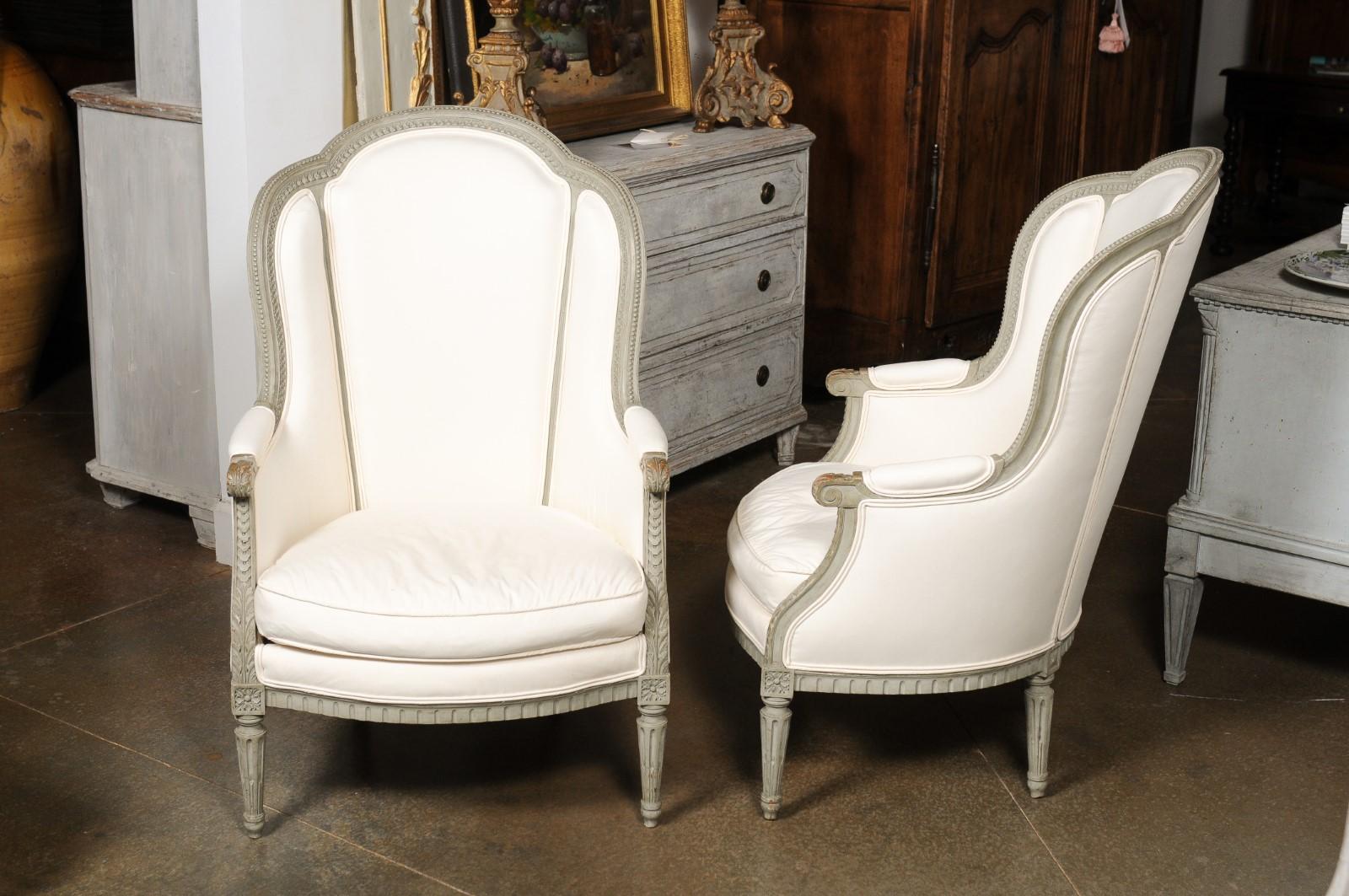 A pair of French Louis XVI style painted wood wingback bergères chairs from the 19th century, with carved décor and new upholstery. Created in France during the 19th century, each of this pair of bergères features a winged back, designed to provide