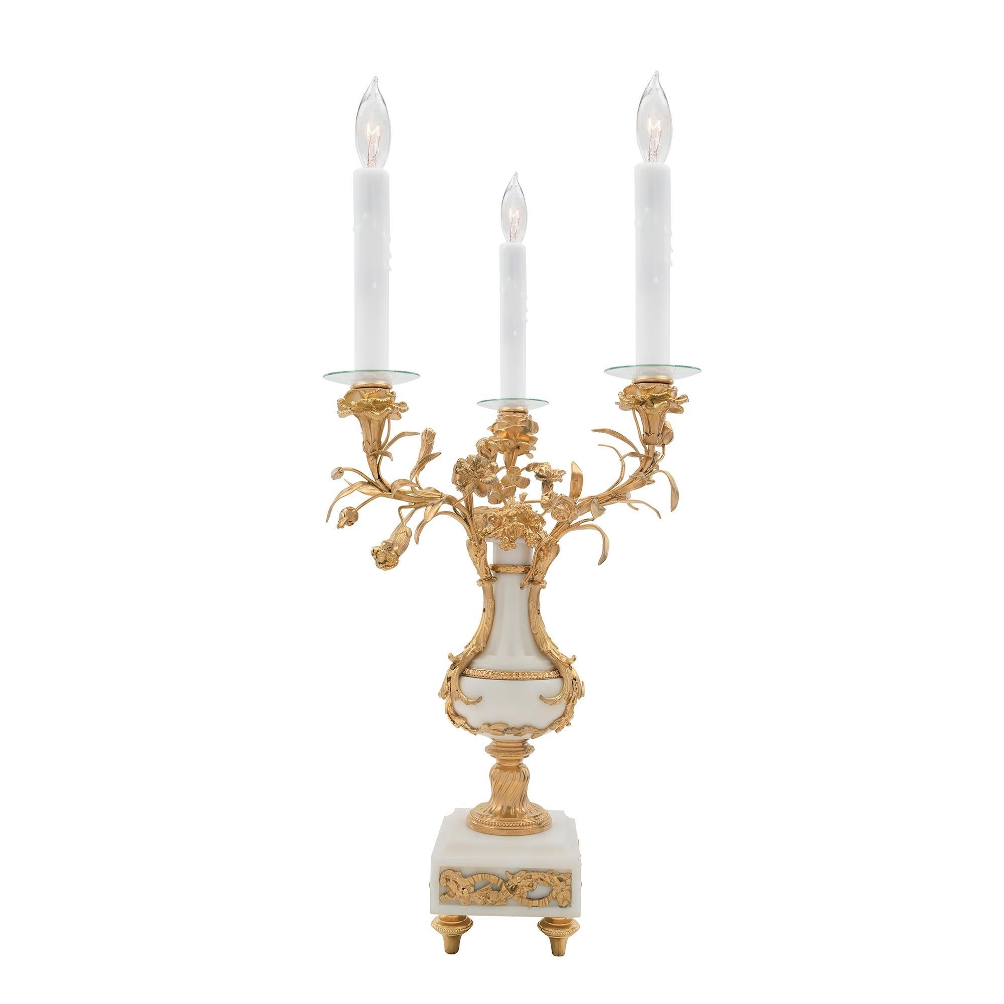 A pair of elegant and uniquely shaped French 19th Century Louis XVI st. three-arm electrified candelabras. Each is raised by a square white carrara marble base with recessed central panels and pierced ormolu mounts all above topie shaped ormolu