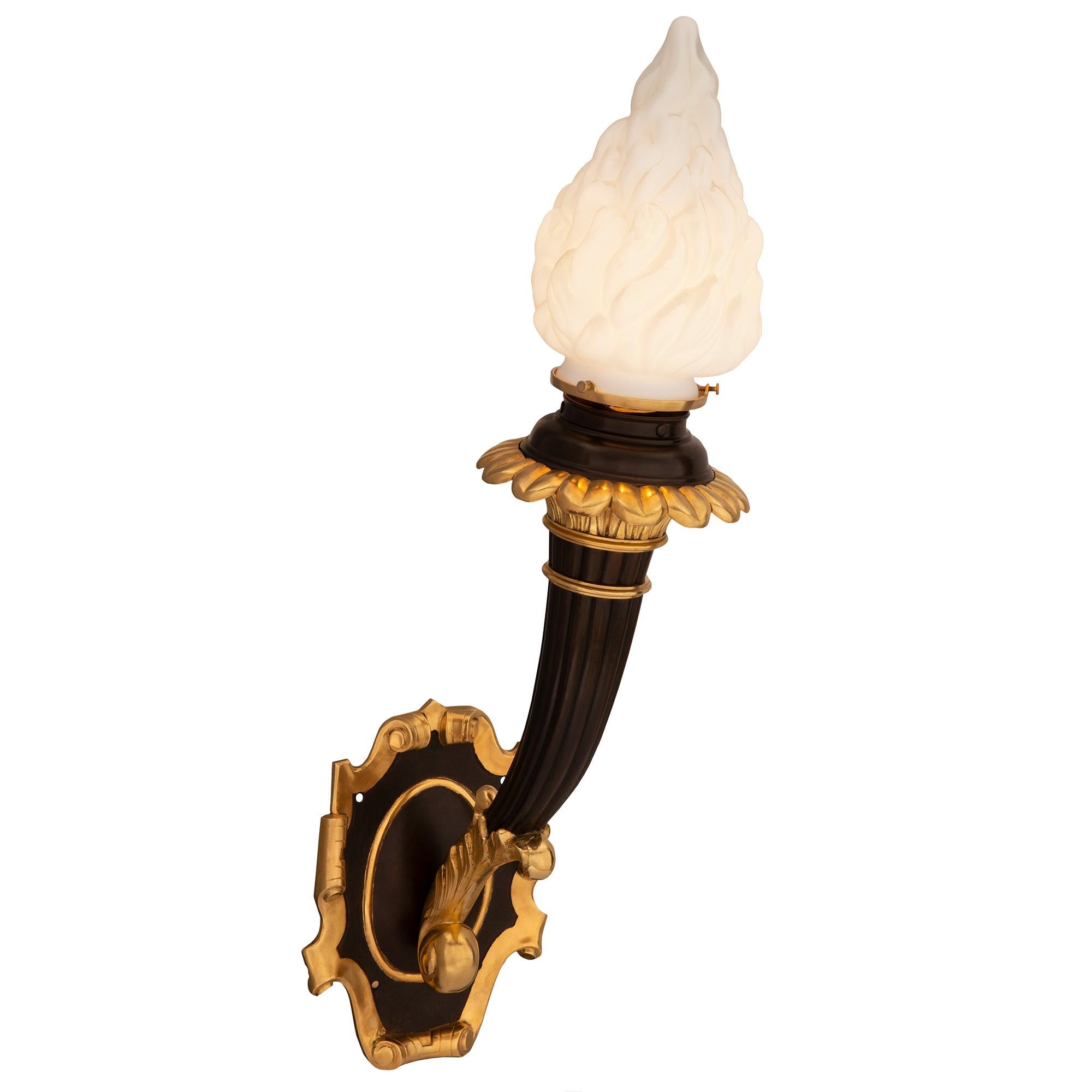 A striking pair of French 19th century Neo-Classical st. patinated bronze and ormolu Bras de Lumière sconces. Each sconce is centered by a beautiful scrolled backplate from where the elegant lightly curved reeded supports extend. The supports
