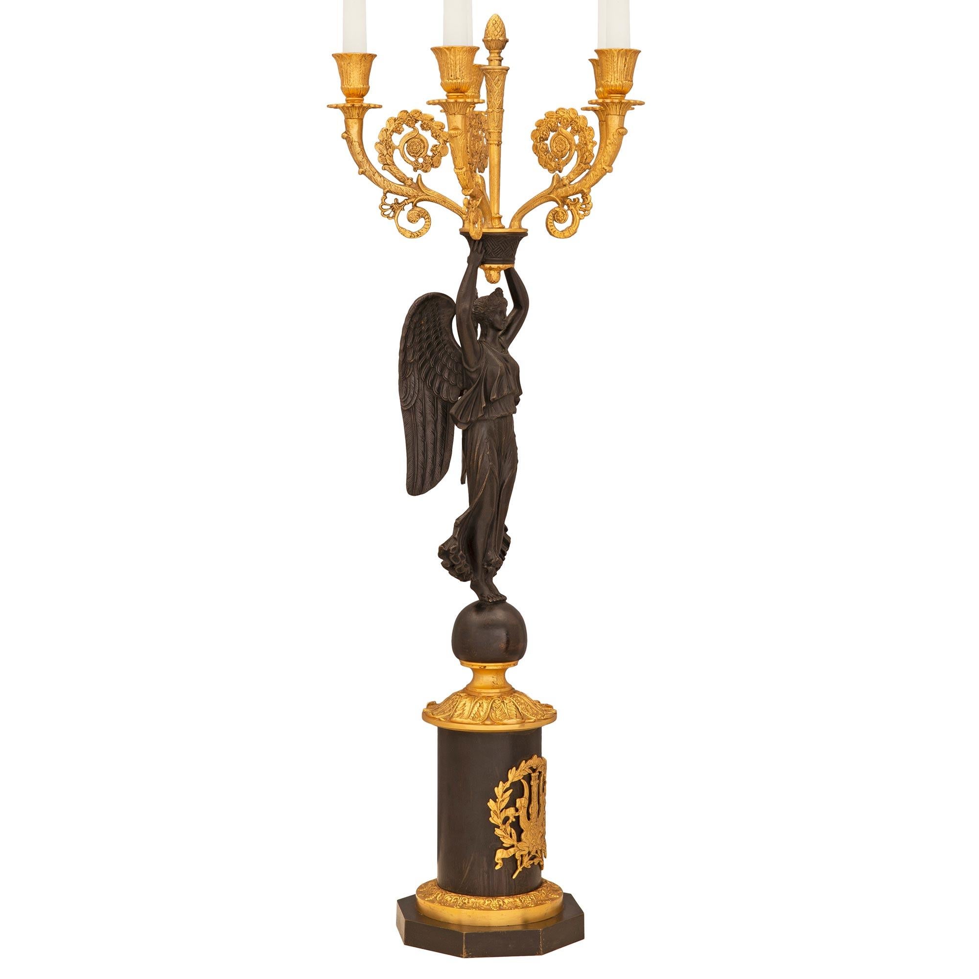 Une très belle et grande paire de candélabres à cinq bras de style néo-classique français du 19ème siècle. La paire en bronze doré et patiné repose sur une base circulaire avec un décor en bronze doré ciselé au centre, sous une figure féminine ailée