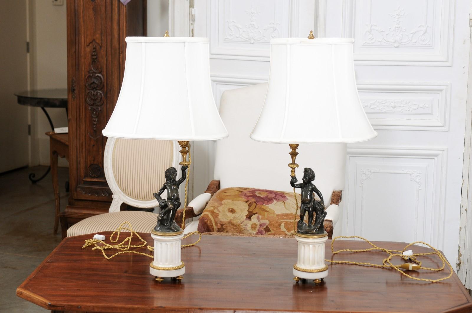 A pair of French neoclassical style white marble and bronze putti lamps from the 19th century, with shades. Created in France during the 19th century, each of this pair of table lamps attracts our attention with its neoclassical decor and