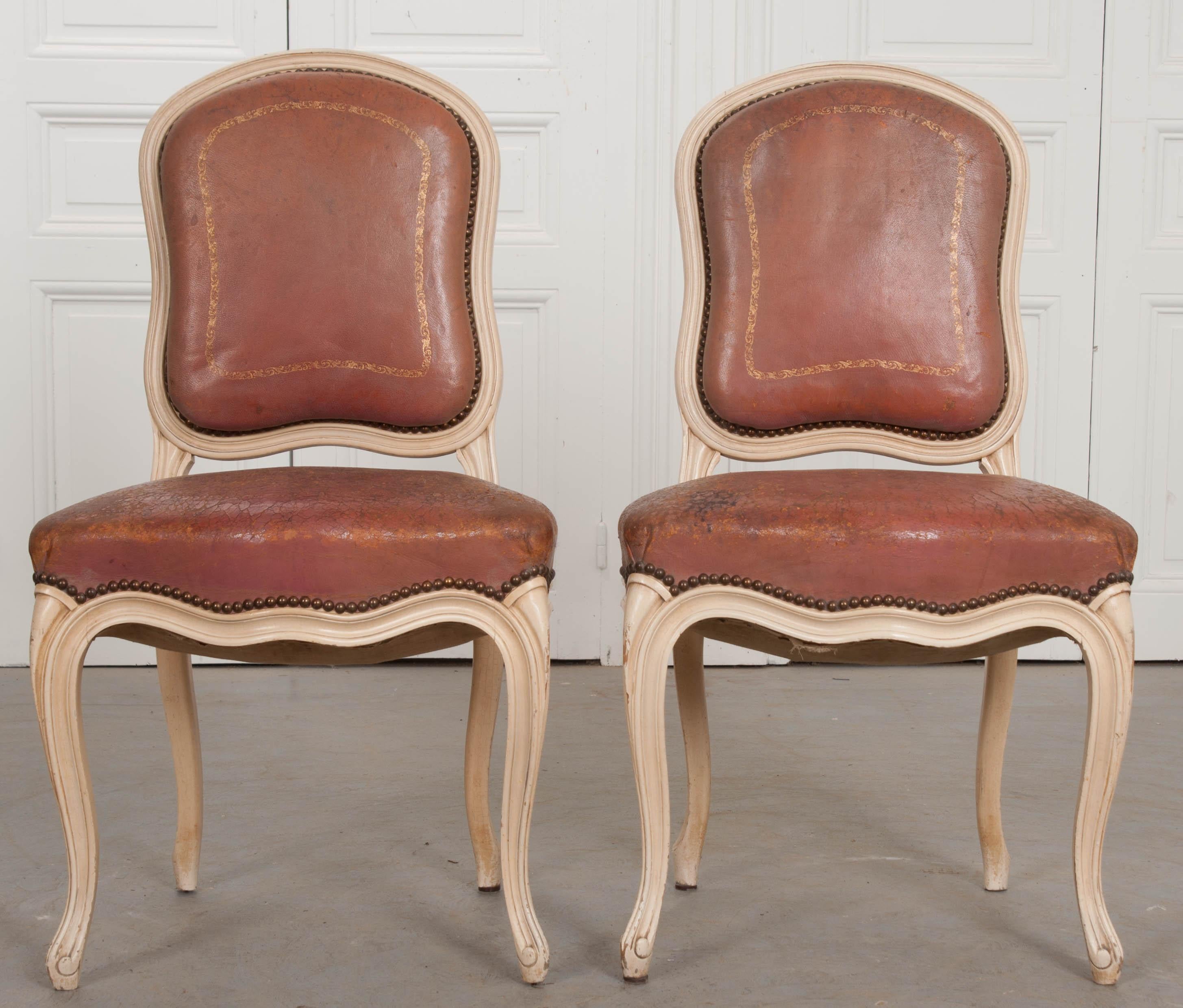 A stunning pair of Louis XV style dining chairs, upholstered in a heavily-worn antique leather. The shapely pair have a frame that is painted in a creamy ivory color that has patinated over time, acquiring an amazing antique finish. They are