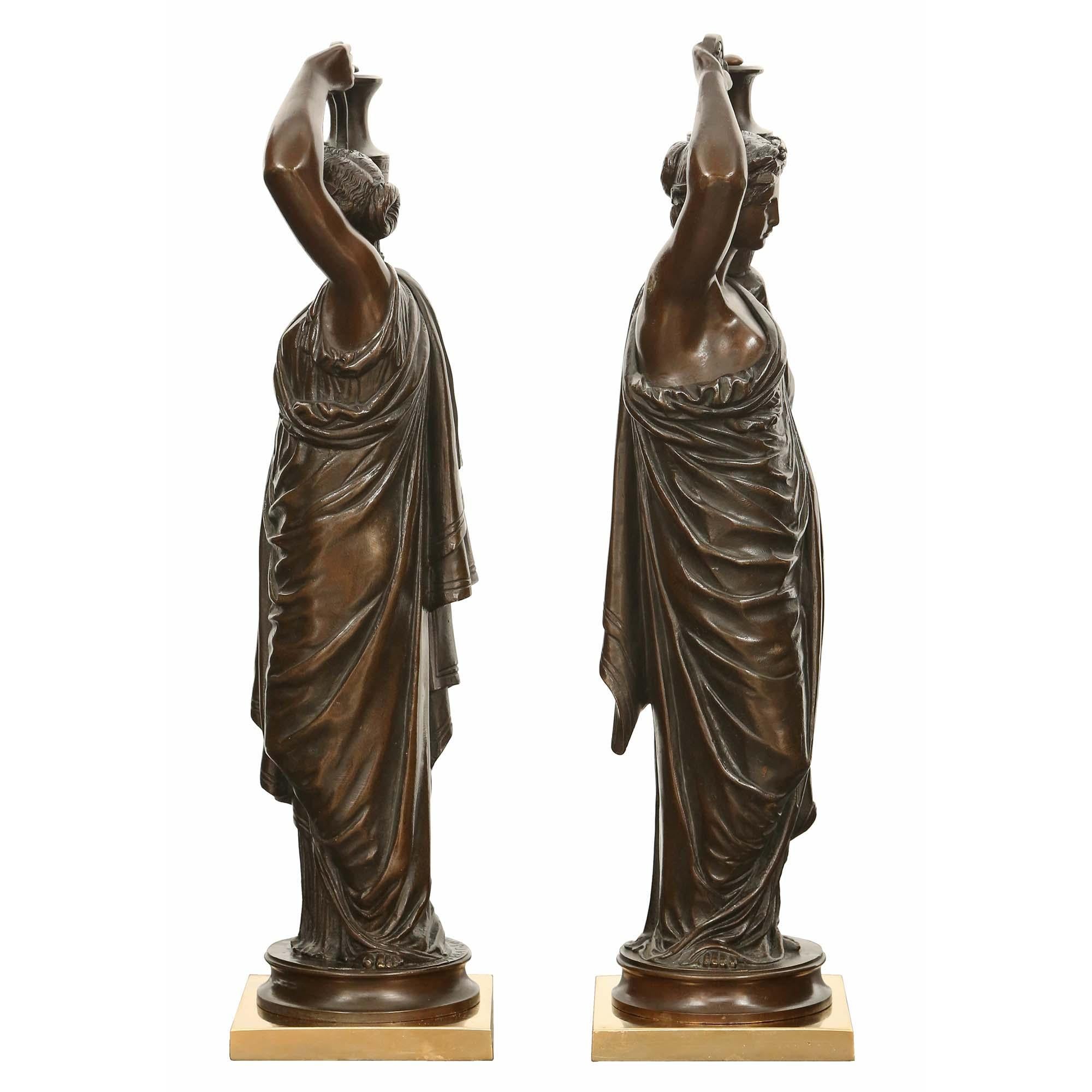 A charming true pair of French 19th century patinated bronze statues signed H. Ferrat. Each bronze is raised by a square ormolu base and with a circular patinated bronze pedestal. The bronzes depict female maidens draped in classical attire carrying