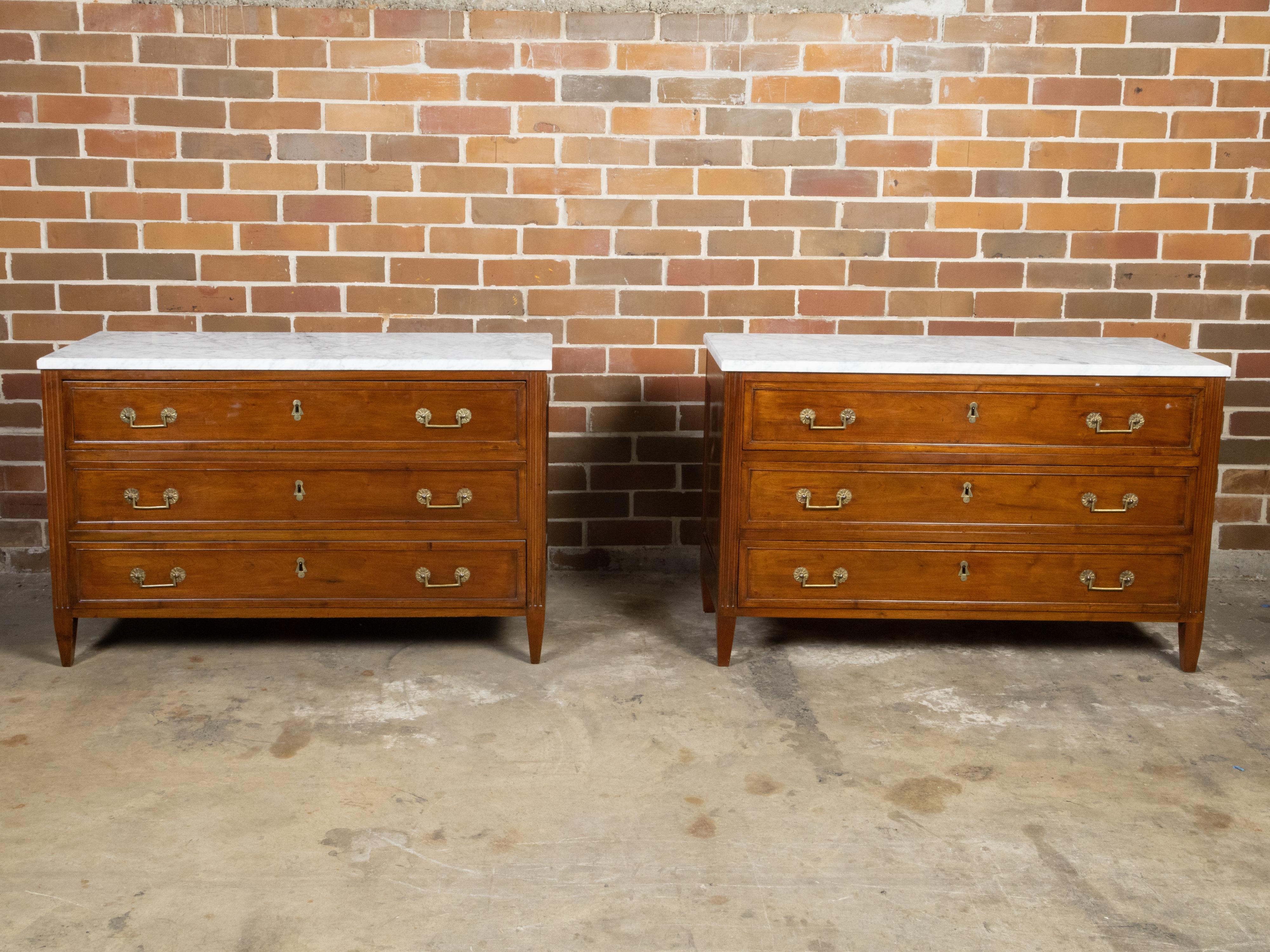 A pair of French Neoclassical style walnut commodes from the 19th century, with white marble tops, three drawers, fluted side posts, tapered legs and brass hardware. Created in France during the 19th century, each of this pair of Neoclassical style