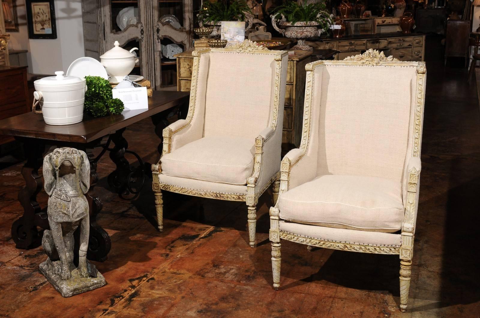 A pair of French Louis XVI style painted and richly carved wingback bergères chairs from the 19th century, re-upholstered in linen. Each of this pair of French 'bergères à oreilles' features an exquisitely carved frame, accented with a delicate