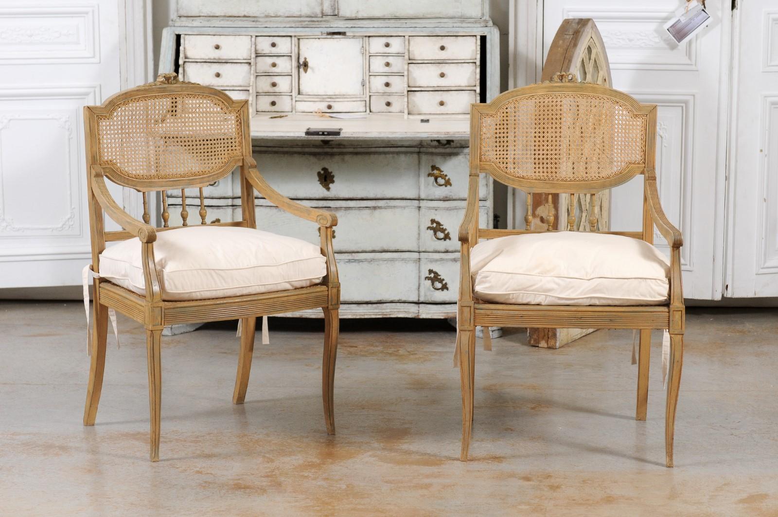 A pair of French wood and cane armchairs from the 19th century, with new custom cushions. Created in France during the 19th century, each of this pair of chairs features a cane back topped with a carved ribbon and adorned with thin spindles in the