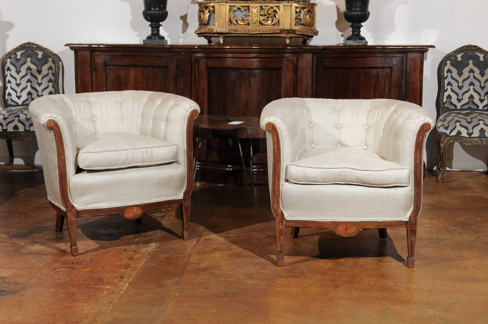 A pair of French wooden club chairs from the 19th century with carved skirt, out-scrolled arms, banded inlay and new upholstery. Each of this pair of French club chairs features a wraparound back, flowing seamlessly into the delicately out-scrolled