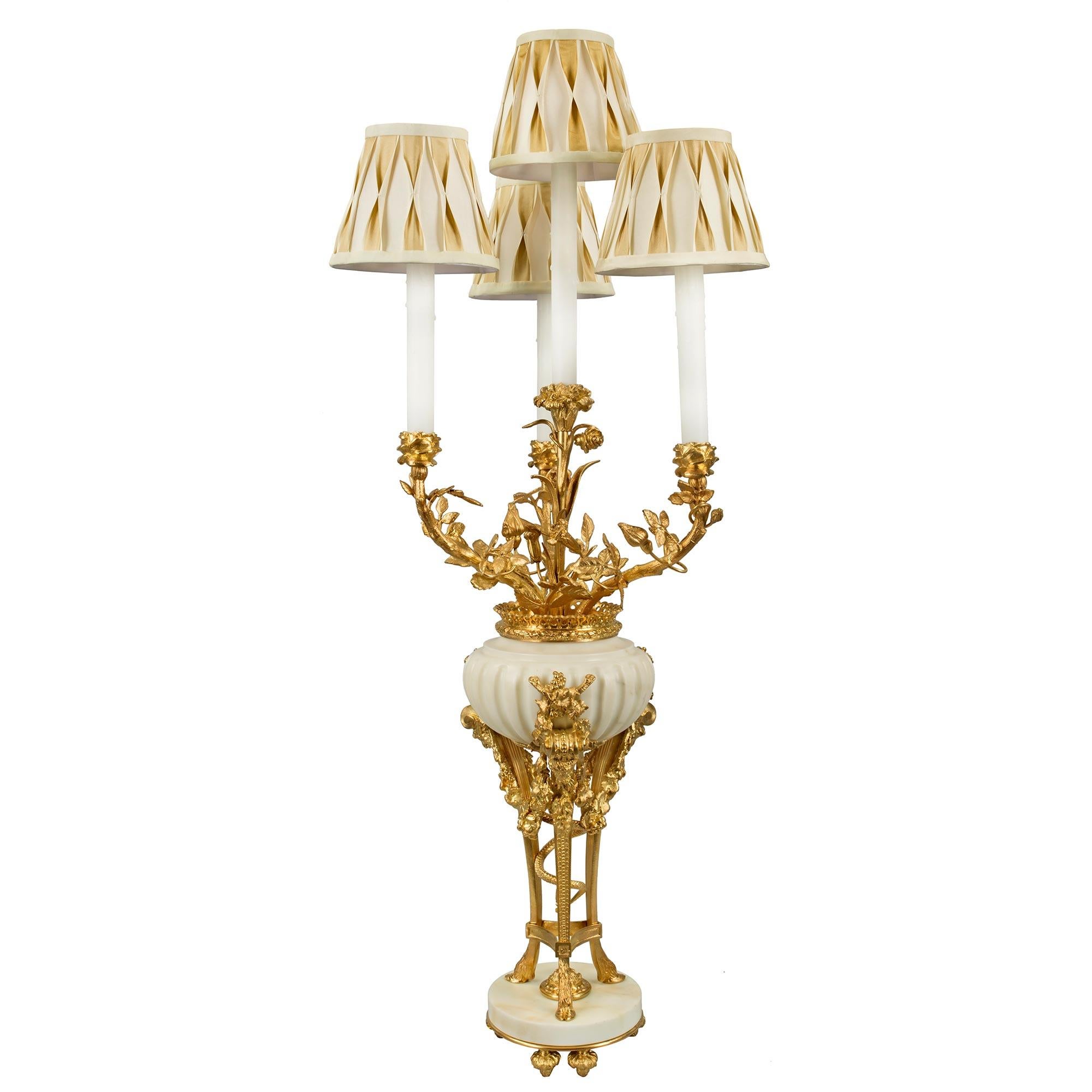 A pair of French 19th Louis XVI st. White Carrara marble and ormolu electrified candelabra lamps, after a model by Pierre Gouthière. Each lamp is raised by ormolu topie shaped feet below a circular marble base. Above is an ormolu tripod with hoof