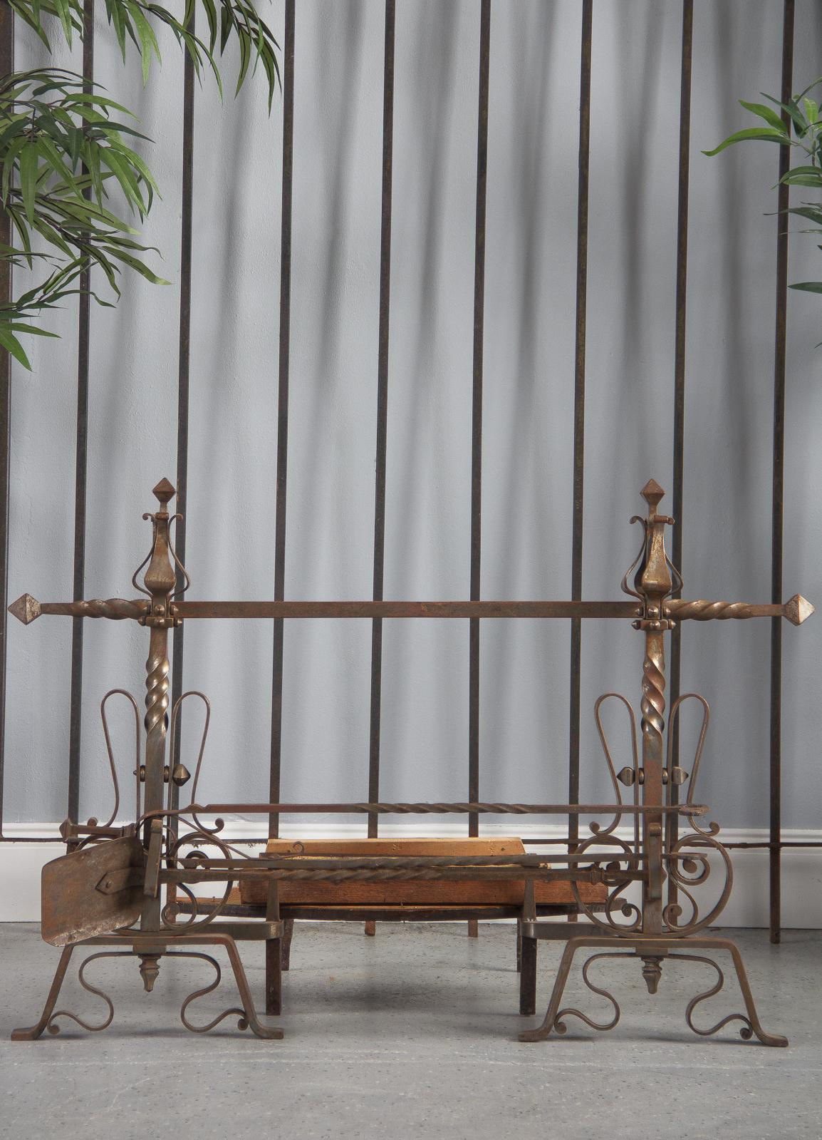 An ornate pair of wrought iron fireplace andirons with guard and accessories, French, 20th century. Heavily scrolled bases sprout posts with twisted elements, capped by curled petal pieces over studded elements, and tipped with square finials. The