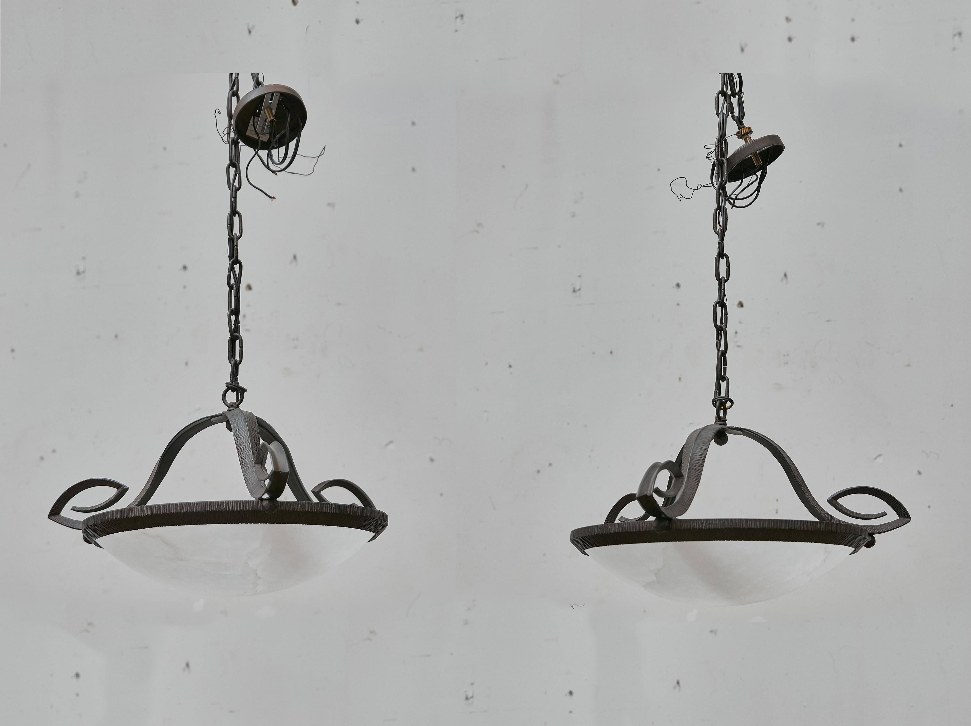 Questa coppia di plafoniere francesi, risalenti al 1930 circa, combina la naturale eleganza dell'alabastro con la forza scultorea del ferro forgiato a mano. Ogni pendente è caratterizzato da una ciotola in alabastro intagliato sospesa all'interno di
